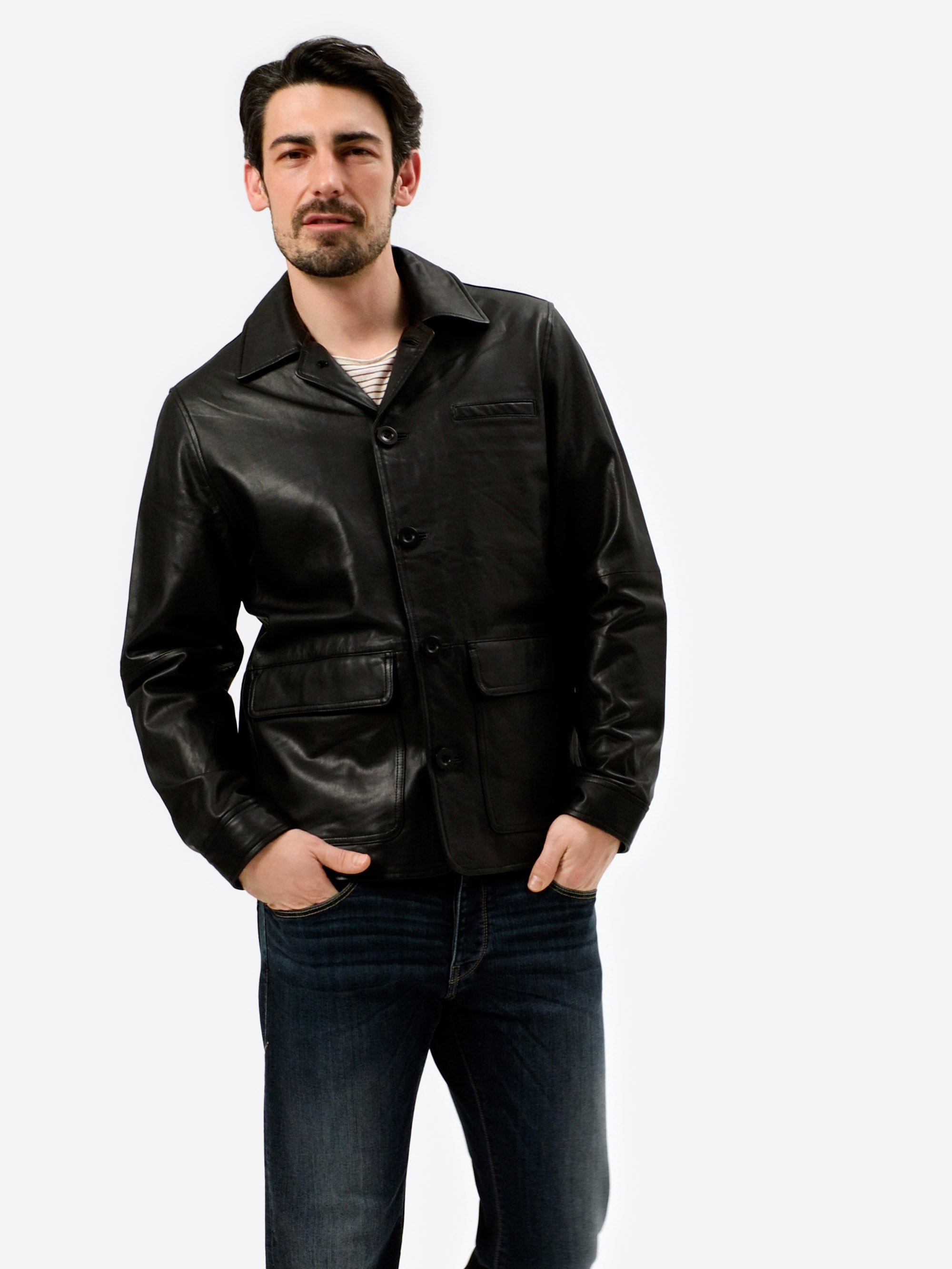 Ein Mann mit dunklem Haar und Bart trägt eine schwarze Lederjacke über einem gestreiften Hemd und blaue Jeans und steht mit einer Hand in der Tasche vor einem schlichten weißen Hintergrund.