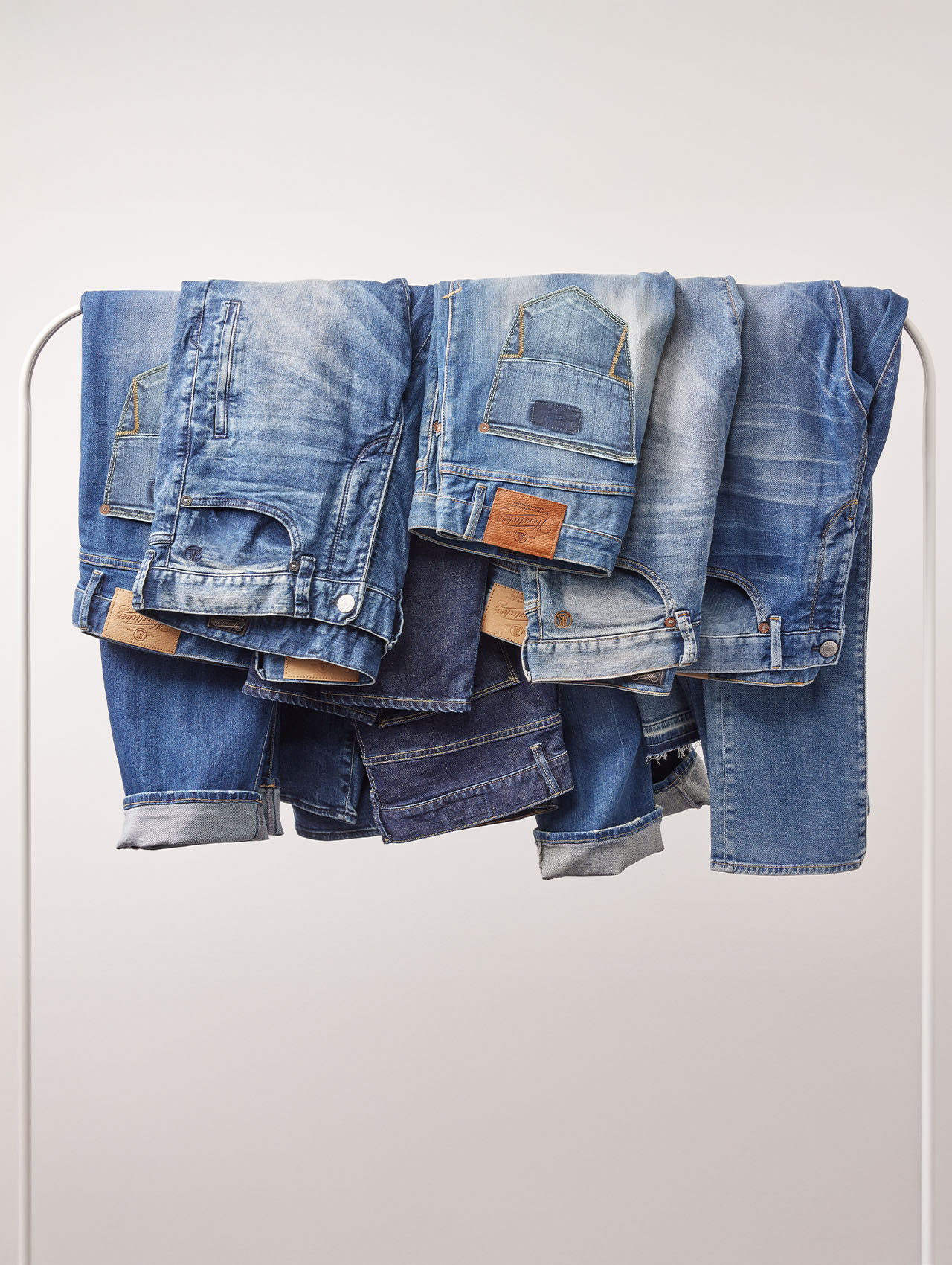 Mehrere blaue Jeans in verschiedenen Waschungen und Stilen sind auf einem weißen Kleiderständer vor einem schlichten, hellen Hintergrund drapiert.