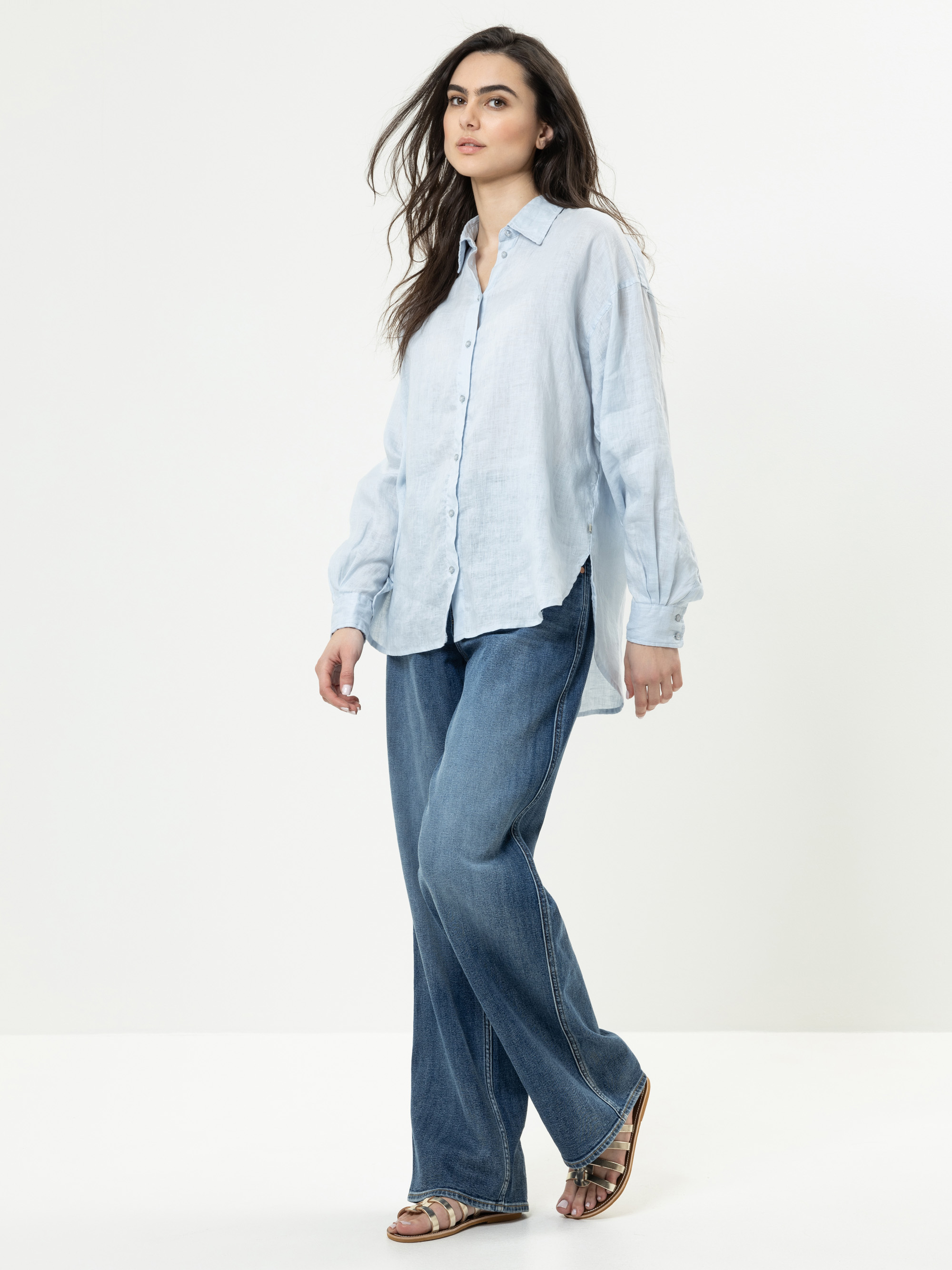 Eine Frau steht vor einem schlichten weißen Hintergrund und trägt ein hellblaues Button-up-Hemd, blaue Jeans mit weitem Bein und Sandalen. Sie hat langes, dunkles Haar und einen neutralen Gesichtsausdruck. Ein Arm liegt entspannt an ihrer Seite, währen