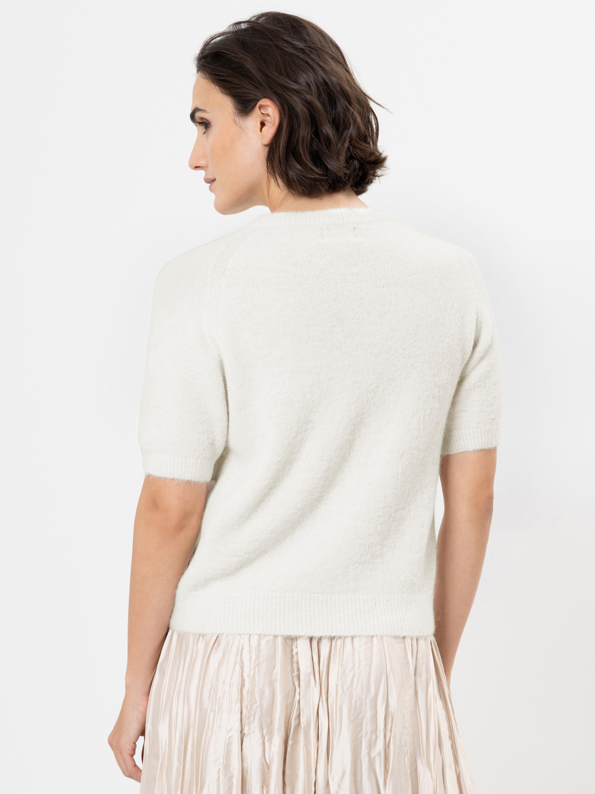 Eine Frau mit kurzen braunen Haaren ist von hinten zu sehen. Sie trägt einen kurzärmeligen, cremefarbenen Pullover und einen leichten Faltenrock vor einem schlichten weißen Hintergrund.