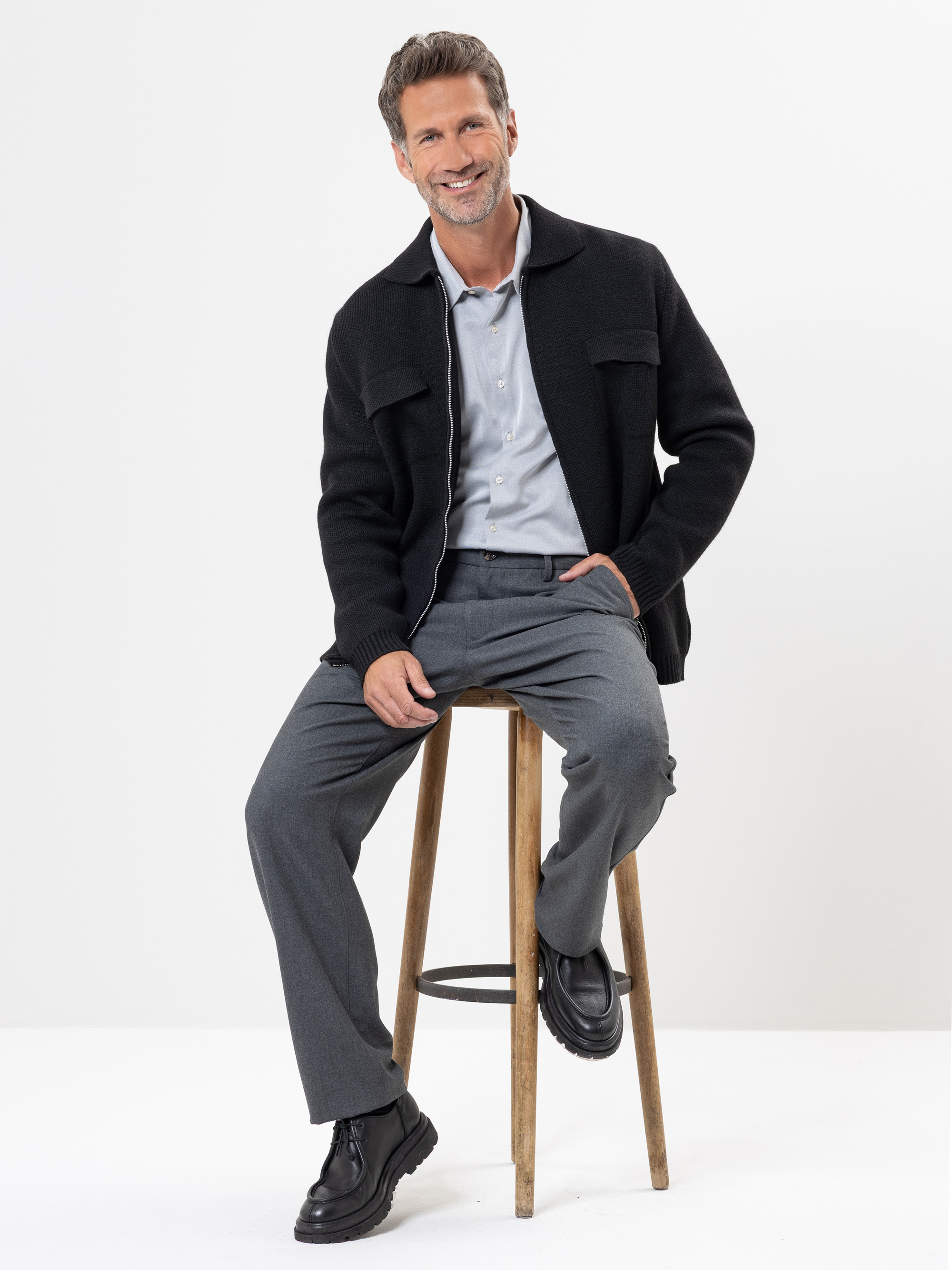 Ein Mann mit kurzem, gestyltem Haar und gestutztem Bart sitzt auf einem Holzschemel und lächelt. Er trägt ein hellblaues Hemd, eine dunkle Jacke, eine graue Hose und schwarze Schuhe vor einem schlichten weißen Hintergrund.