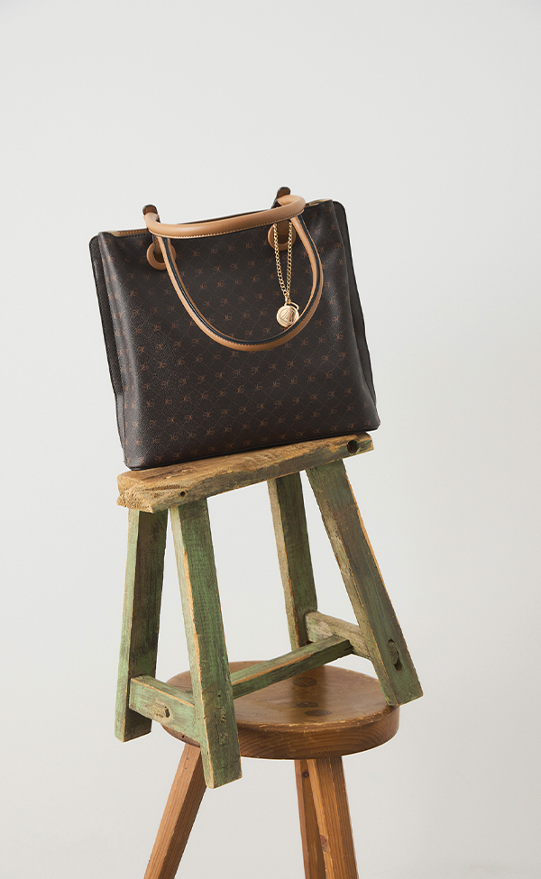 Eine dunkelbraun gemusterte Handtasche mit hellbraunen Griffen und einem goldenen Schlüsselbund sitzt auf einem rustikalen grünen Holzhocker vor einem schlichten weißen Hintergrund.