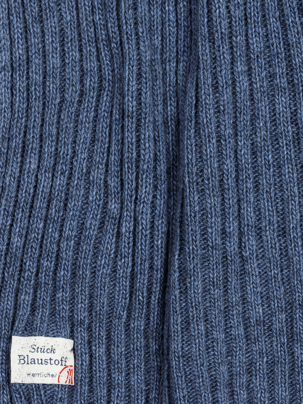 Nahaufnahme eines blauen Rippenstrickstoffs mit einem kleinen weißen Etikett in der unteren linken Ecke, das einen deutschen Text und ein rotes Logo enthält.
