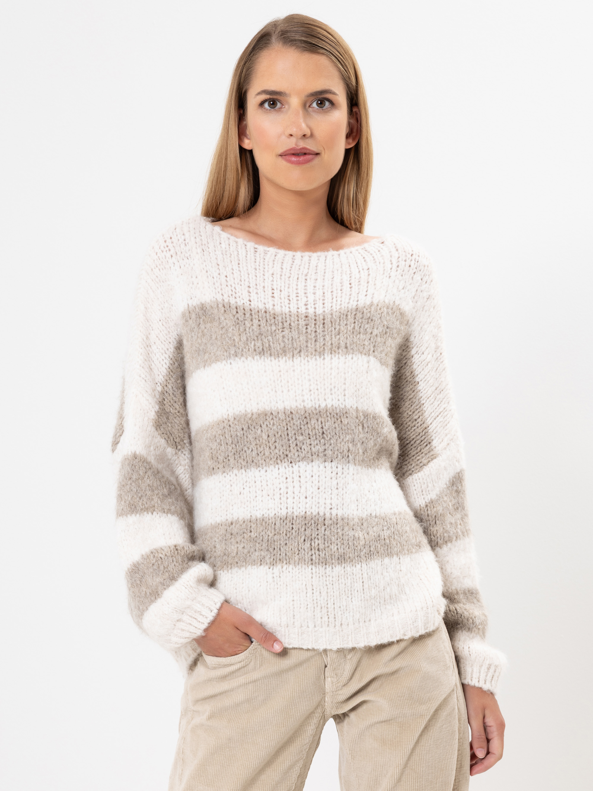 Eine Frau mit glattem, hellbraunem Haar trägt einen gemütlichen, langärmeligen Pullover mit breiten beigen und weißen Querstreifen und eine hellbeige Hose. Sie steht vor einem schlichten weißen Hintergrund und hat eine Hand in der Tasche.