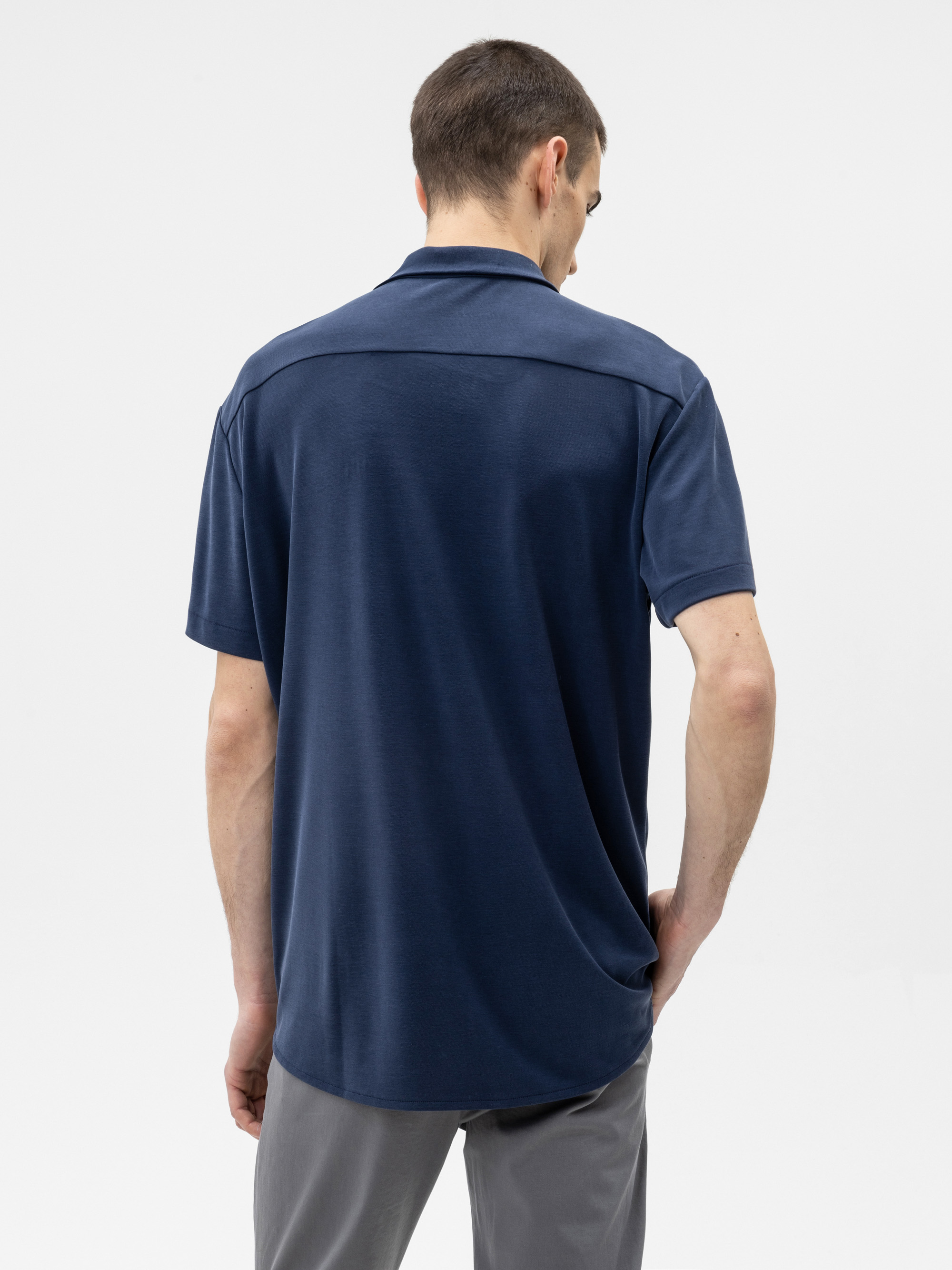 Ein Mann steht von der Kamera abgewandt in einem kurzärmeligen marineblauen Polohemd und einer grauen Hose vor einem schlichten weißen Hintergrund.