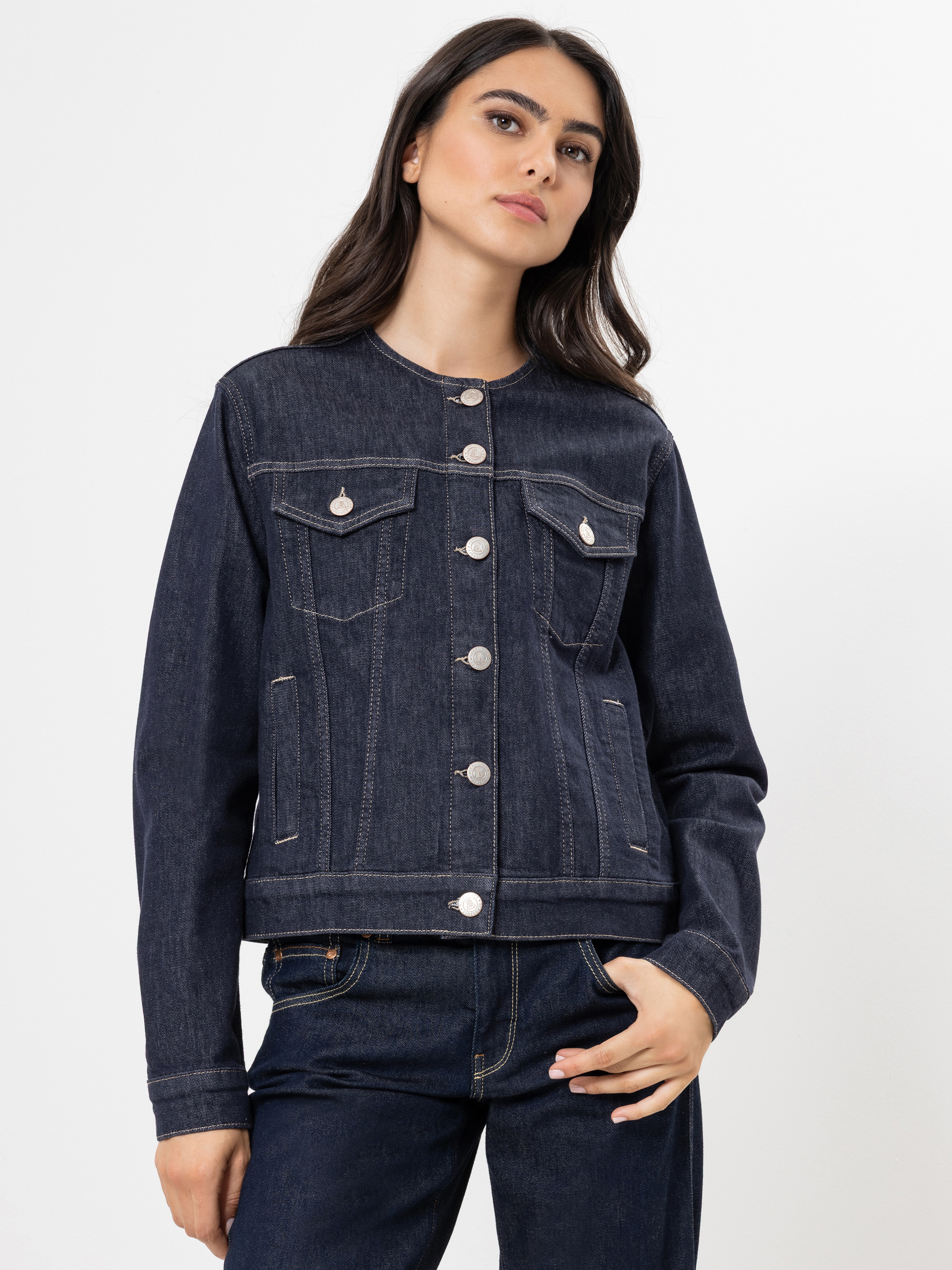 Eine Frau mit langen dunklen Haaren trägt eine dunkelblaue, hochgeknöpfte Jeansjacke und dazu passende Jeans, die vor einem schlichten weißen Hintergrund stehen. Sie blickt selbstbewusst in die Kamera und hat eine Hand in der Tasche.