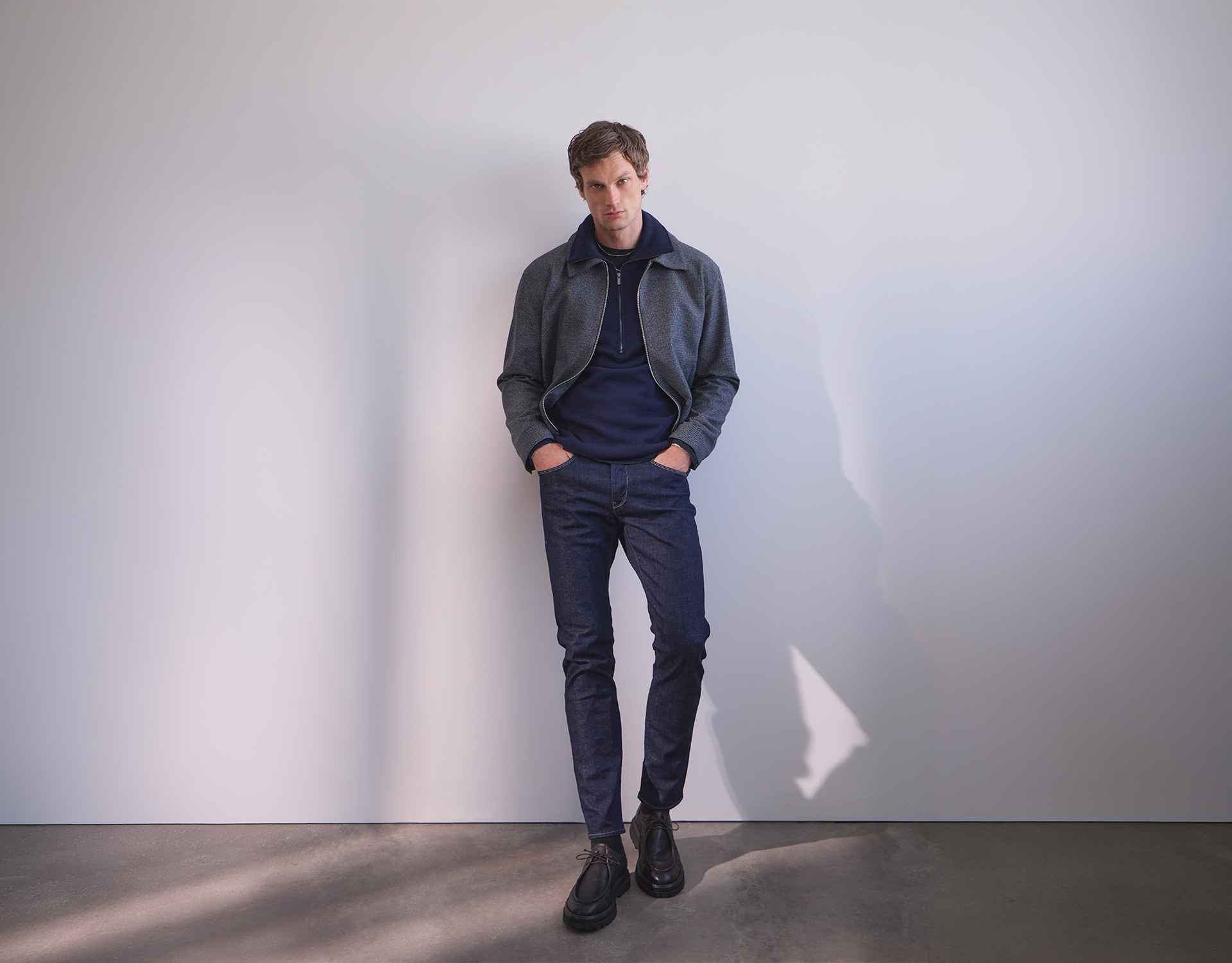 Ein Mann steht mit den Händen in den Taschen vor einer schlichten, hellen Wand. Er trägt eine dunkle Jacke, einen marineblauen Pullover, blaue Jeans und schwarze Schuhe. Der Boden ist grau, und sein Schatten ist an der Wand zu sehen.