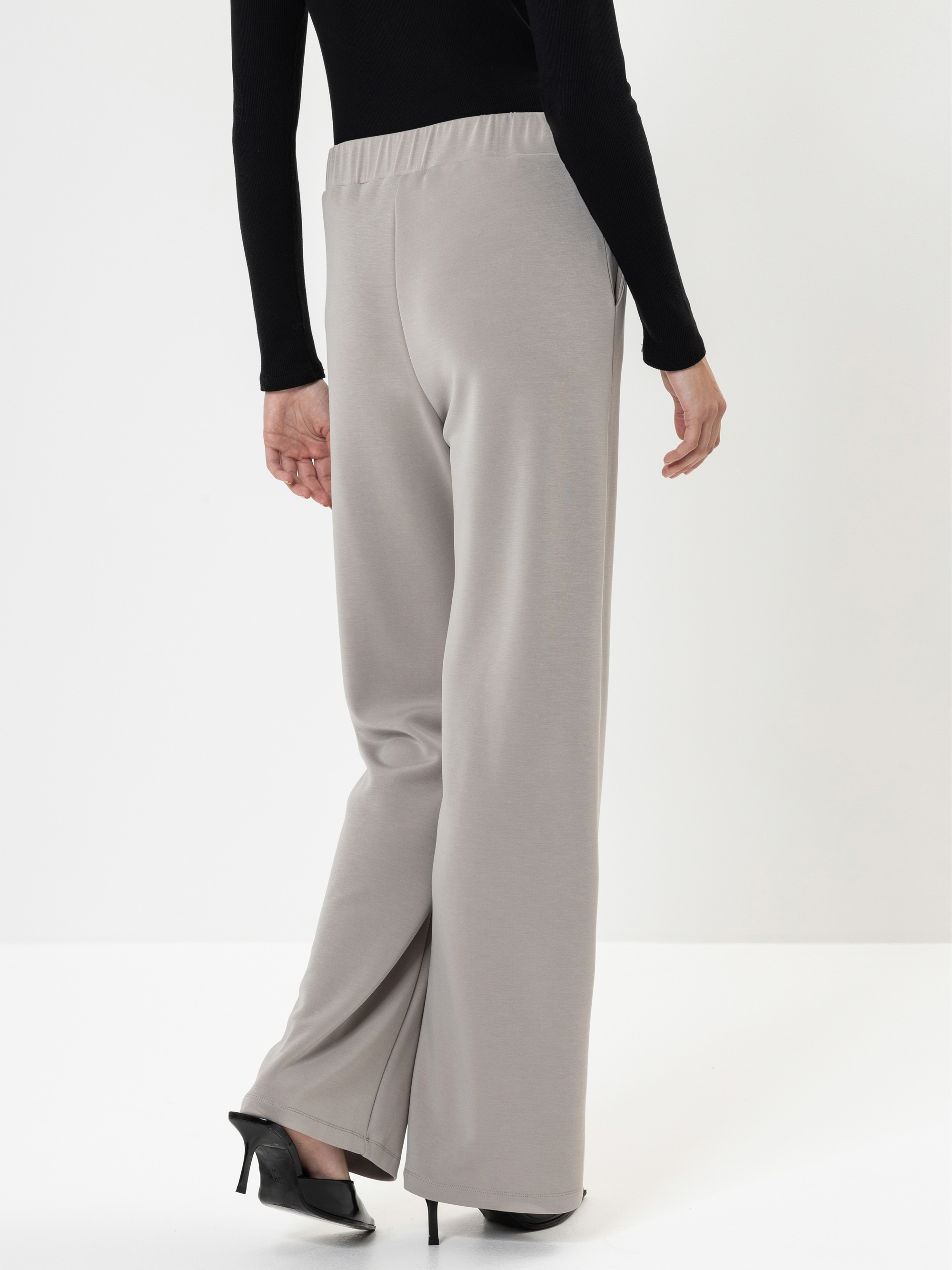 Eine Person, die eine hellgraue Hose mit weitem Bein, ein schwarzes langärmeliges Oberteil und schwarze hochhackige Schuhe trägt, steht vor einem schlichten weißen Hintergrund und blickt von der Kamera weg.