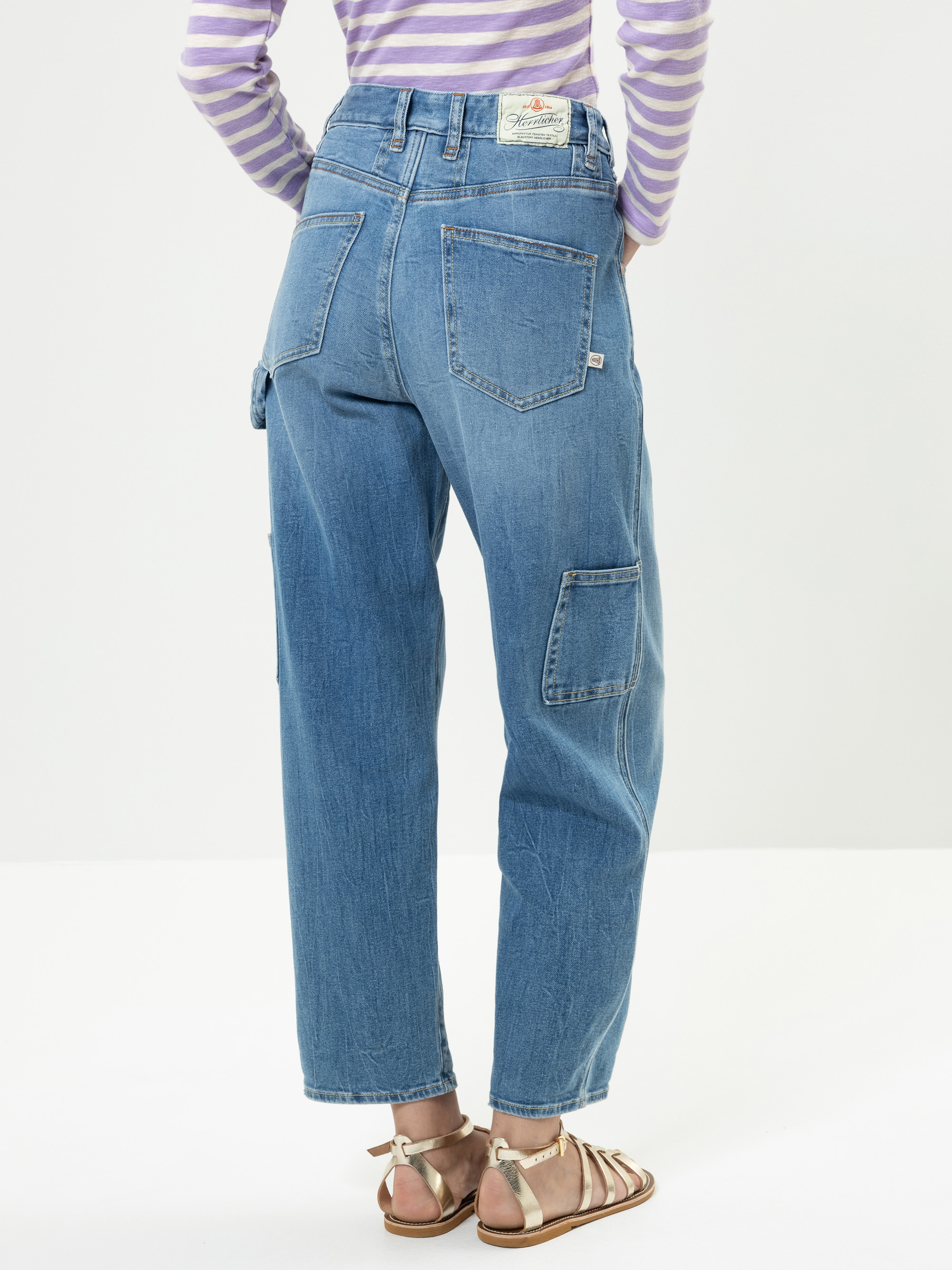 Eine Person steht mit dem Gesicht zur Wand und trägt eine weite blaue Jeans mit großen Gesäßtaschen und ein lila-weiß gestreiftes langärmeliges Hemd. Außerdem trägt sie flache Riemchensandalen auf einem schlichten weißen Hintergrund.