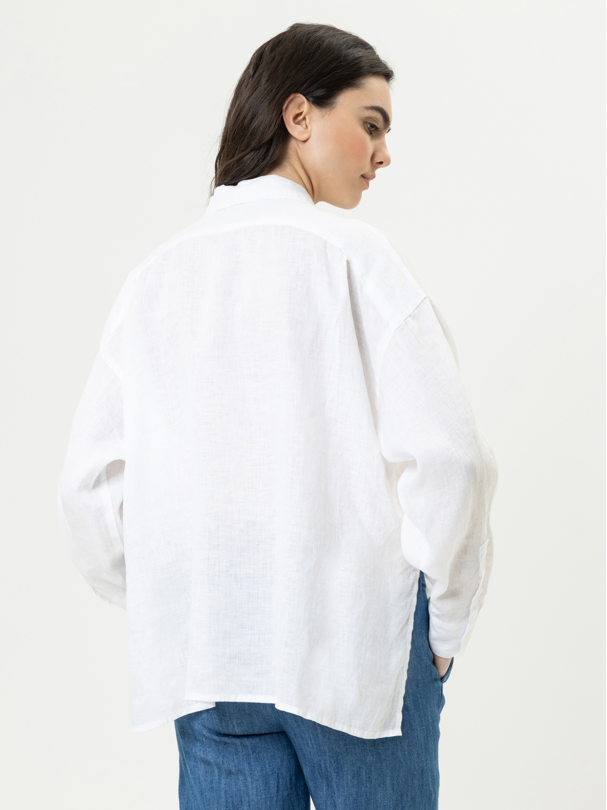 Eine Frau mit langen dunklen Haaren ist von hinten zu sehen. Sie trägt ein locker sitzendes weißes Button-up-Hemd und blaue Jeans und steht vor einem schlichten hellen Hintergrund.