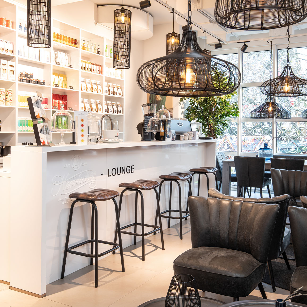 Modernes Café-Interieur mit weißem Tresen, vier hohen Hockern, hängenden schwarzen Drahtlampen, Regalen mit Produkten, Kaffeemaschinen und stilvollen schwarzen Stühlen. Große Fenster lassen natürliches Licht herein, und der Raum ist mit Grünpflanze