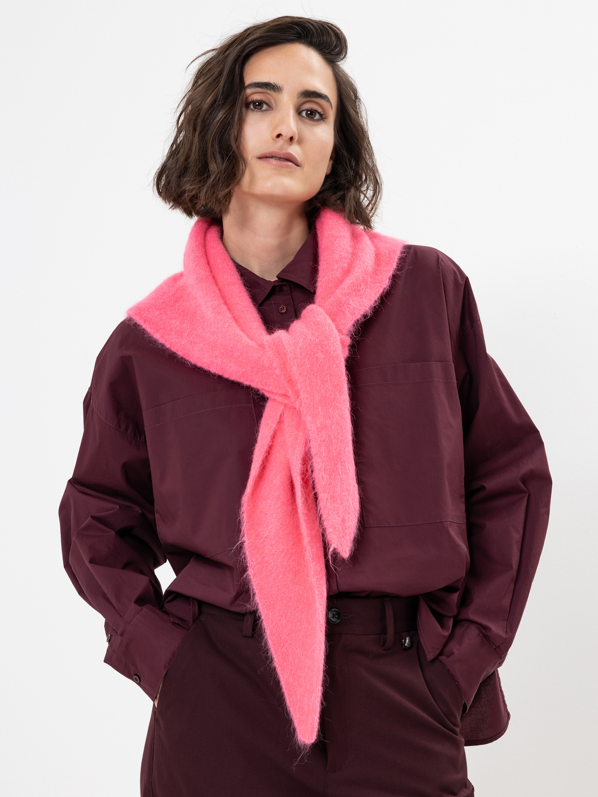 Eine Frau steht vor einem weißen Hintergrund, trägt ein weinrotes Hemd und eine Hose und hat einen hellrosa, wuscheligen Schal um den Hals gelegt. Sie hat gewelltes, schulterlanges dunkles Haar und einen neutralen Gesichtsausdruck.