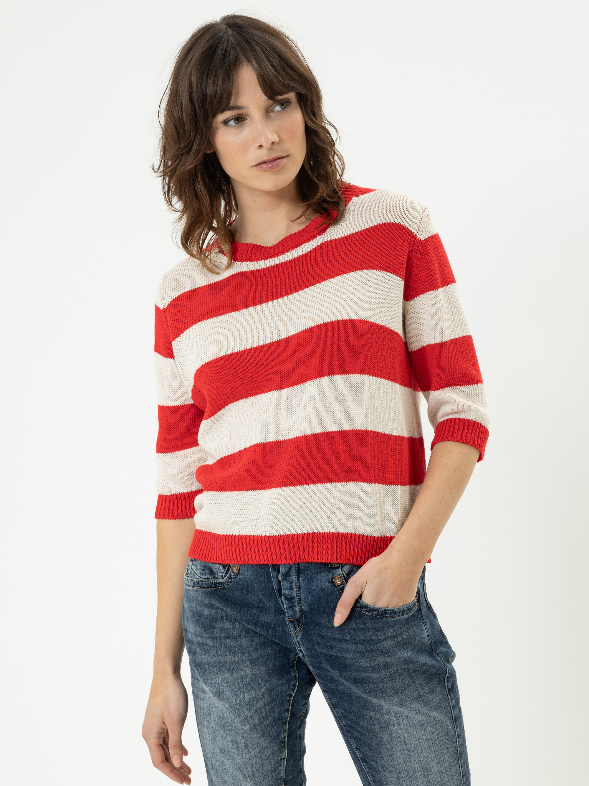 Eine Frau mit gewelltem braunem Haar, die einen rot-weiß gestreiften Pullover und blaue Jeans trägt, steht mit einer Hand in der Hosentasche und schaut leicht zur Seite vor einem schlichten weißen Hintergrund.