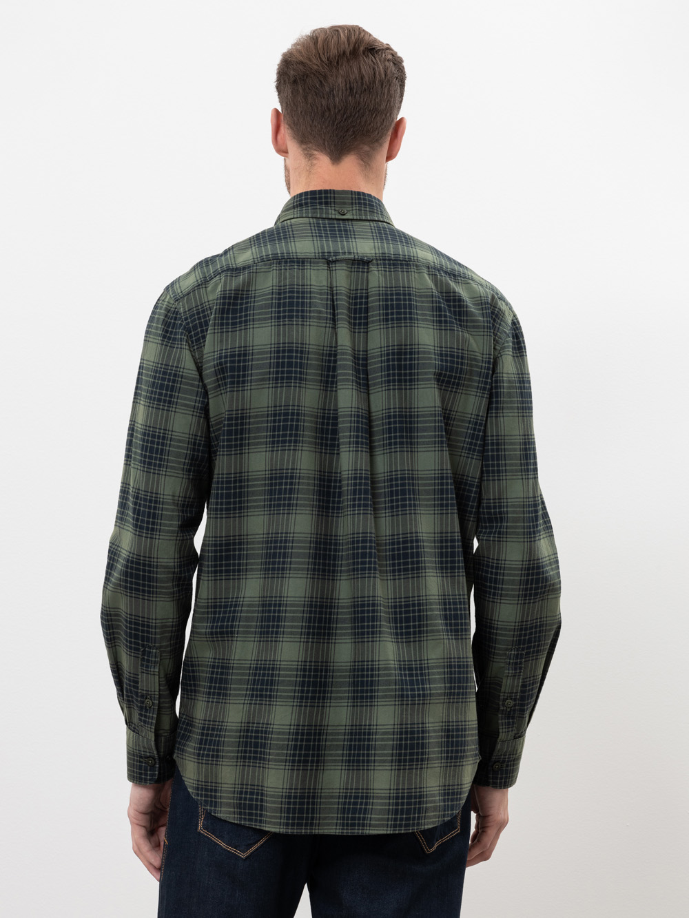 Ein Mann steht mit einem grün und marineblau karierten langärmeligen Hemd und einer dunkelblauen Jeans vor einem schlichten weißen Hintergrund und blickt von der Kamera weg.