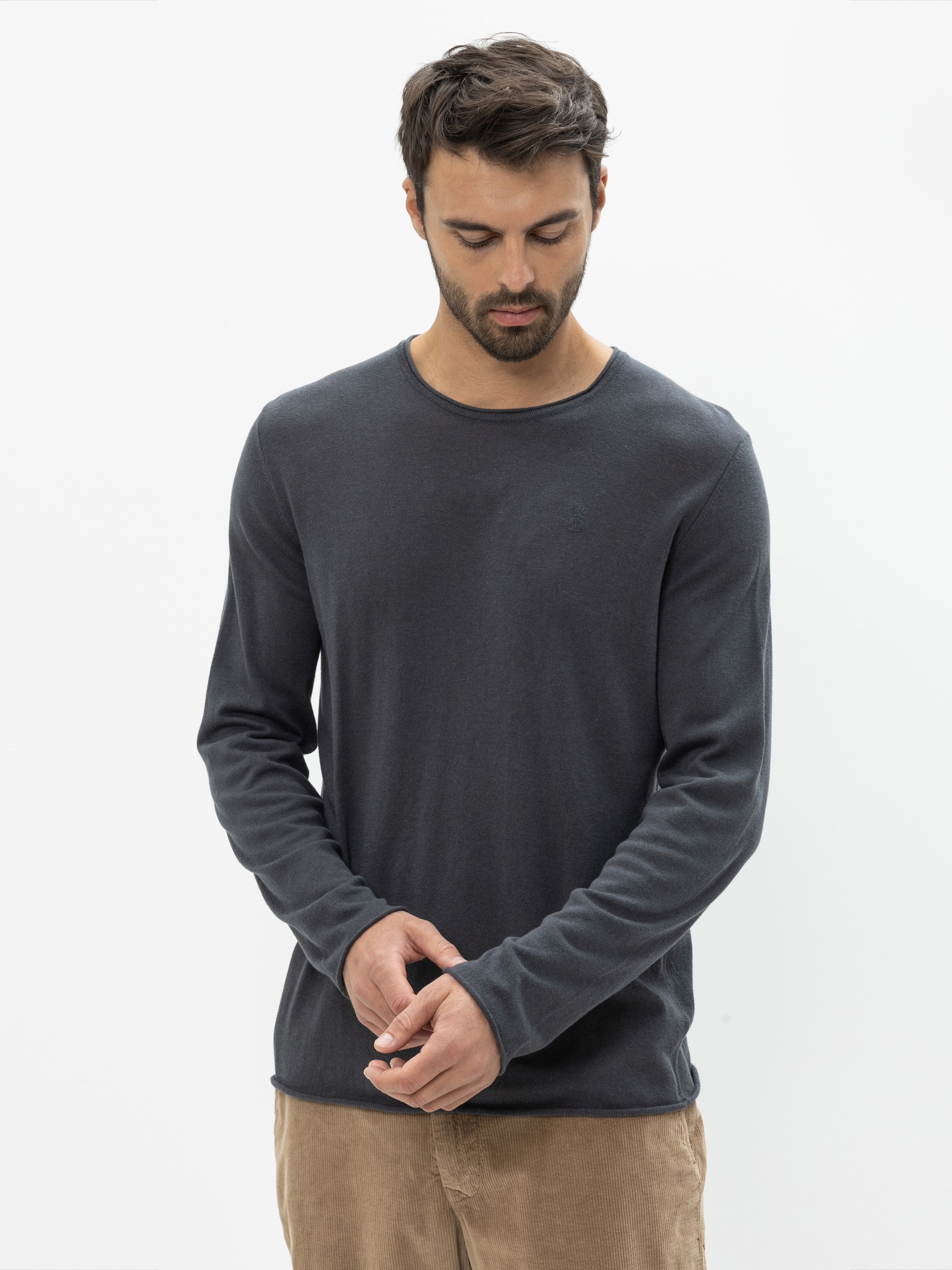 Ein Mann mit kurzen dunklen Haaren und einem Bart trägt ein dunkles langärmeliges Hemd und eine beigefarbene Hose. Er steht vor einem schlichten weißen Hintergrund und blickt nach unten, während er seinen Ärmel zurechtrückt.