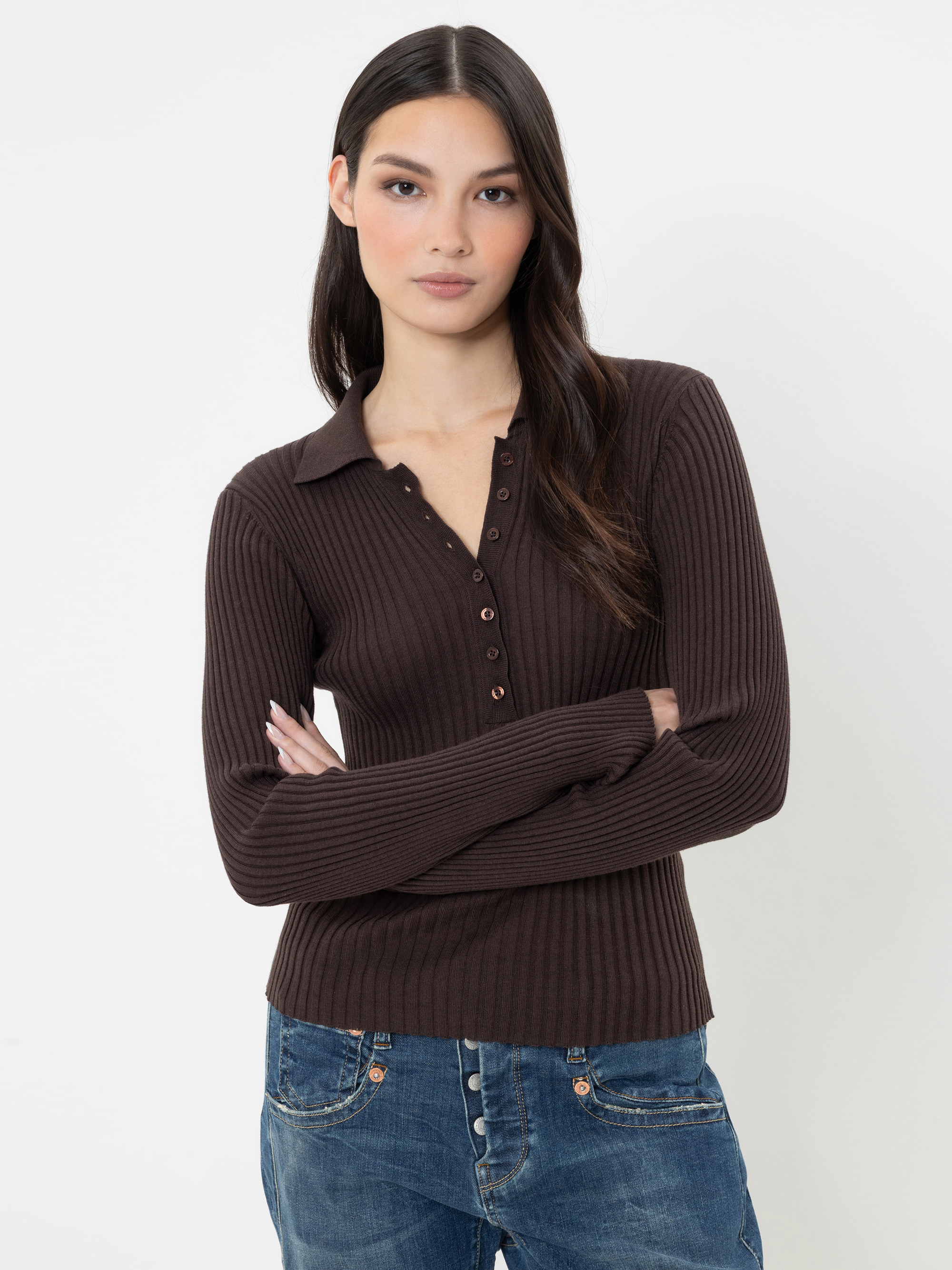 Eine Frau mit langen dunklen Haaren, die einen gerippten, dunkelbraunen Pullover mit Knöpfen und eine blaue Jeans trägt, steht mit verschränkten Armen vor einem schlichten weißen Hintergrund.