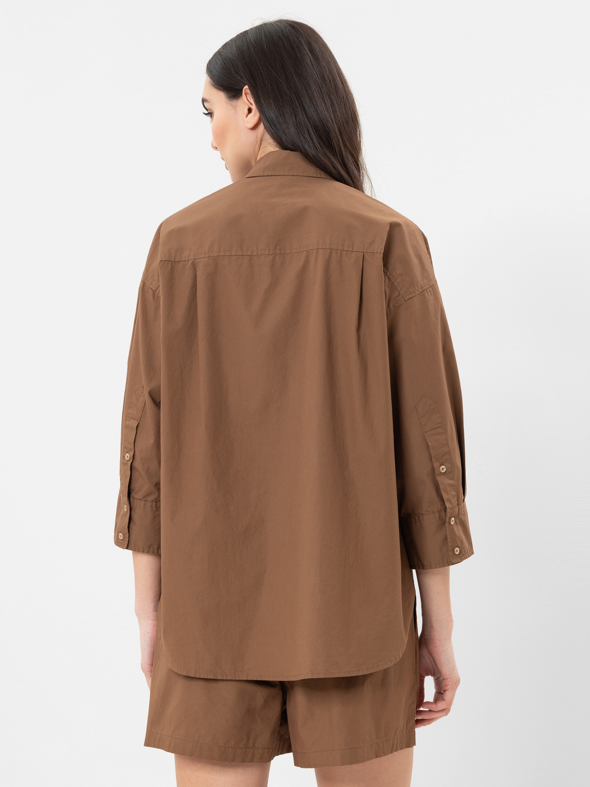 Eine Frau mit langen braunen Haaren, von hinten gesehen, trägt ein locker sitzendes braunes Button-up-Hemd mit gekrempelten Ärmeln und passende braune Shorts vor einem schlichten weißen Hintergrund.