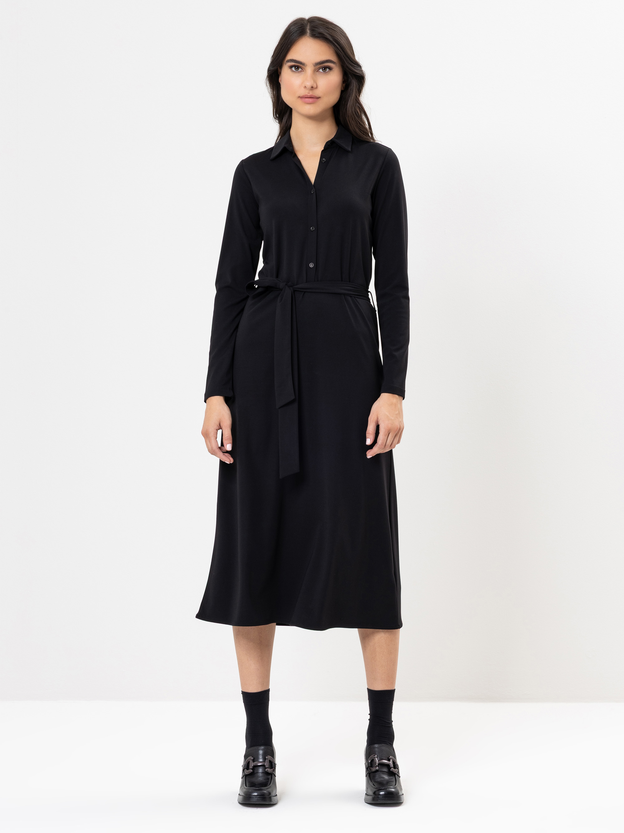 Eine Frau steht vor einem schlichten weißen Hintergrund und trägt ein langärmeliges, hochgeknöpftes schwarzes Midikleid mit passender Krawatte, schwarzen Socken und schwarzen Halbschuhen.
