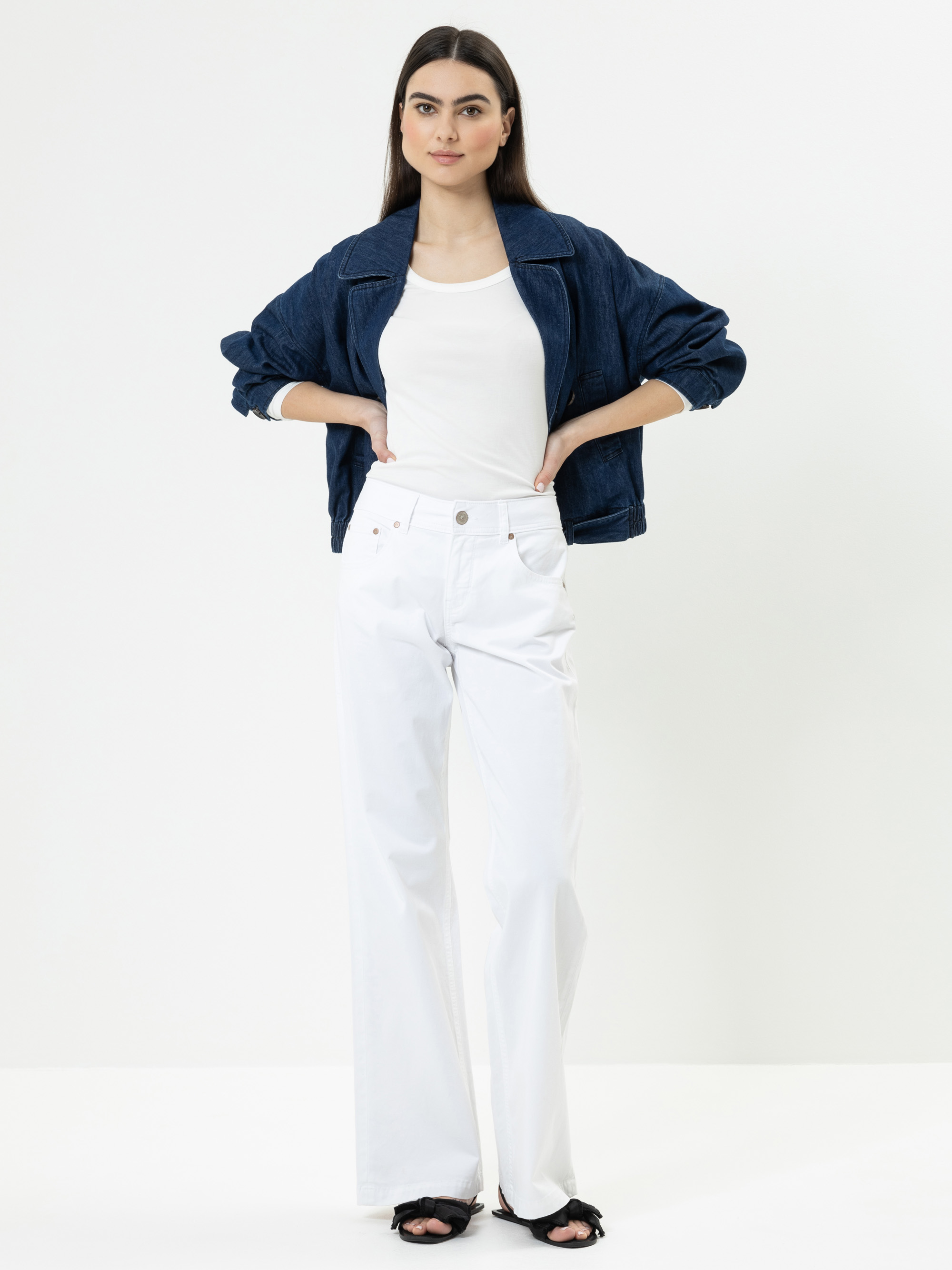 Eine Frau steht vor einem weißen Hintergrund und trägt eine dunkelblaue Jeansjacke über einem weißen Oberteil, eine weiße Hose mit weitem Bein und schwarze Sandalen, wobei sie ihre Hände in die Taille stützt.