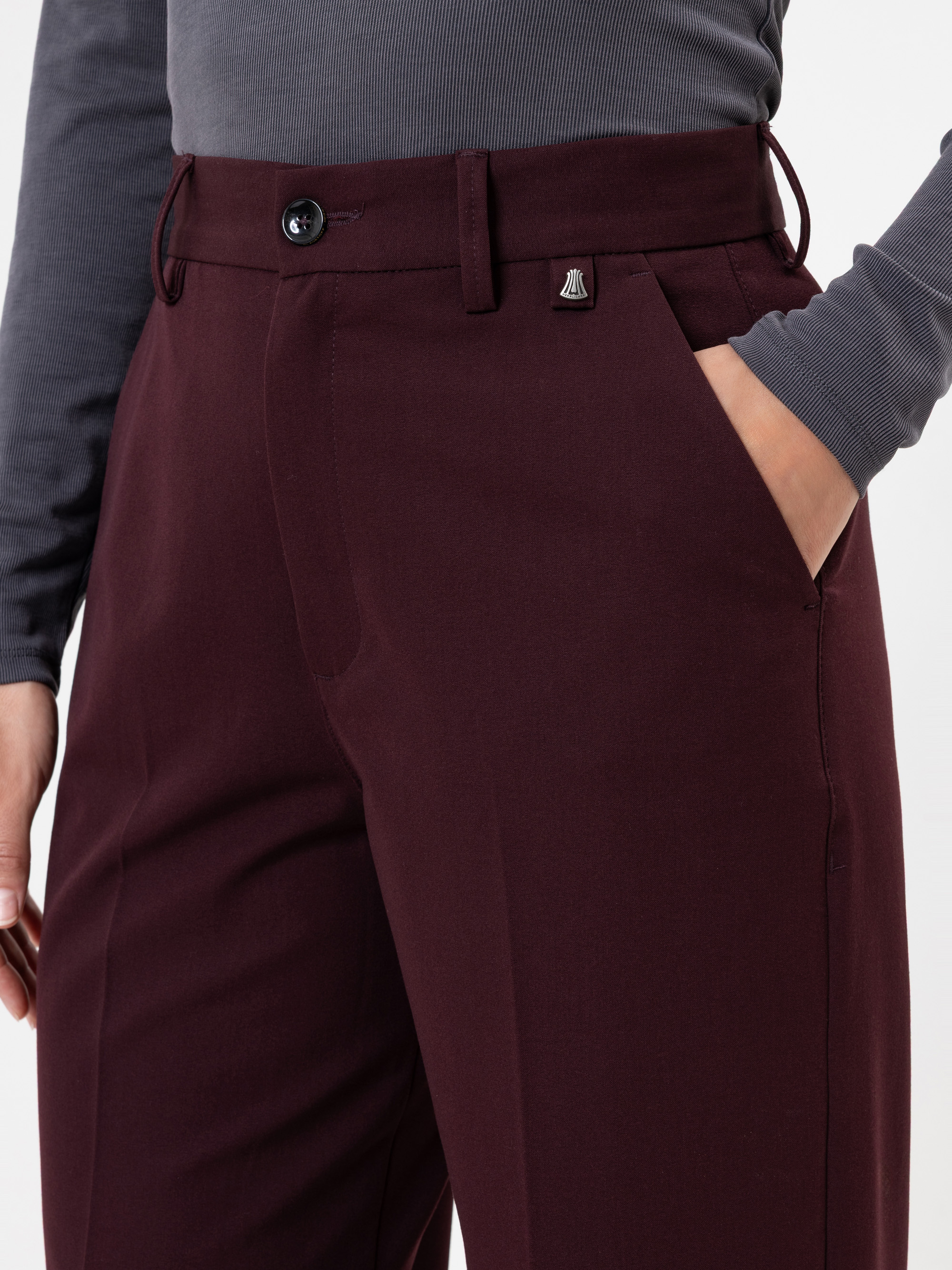 Eine Person, die eine hochtaillierte, weinrote Hose mit Knopf und Reißverschluss und ein langärmeliges, tailliertes graues Oberteil trägt. Ihre Hand befindet sich in einer der Hosentaschen.