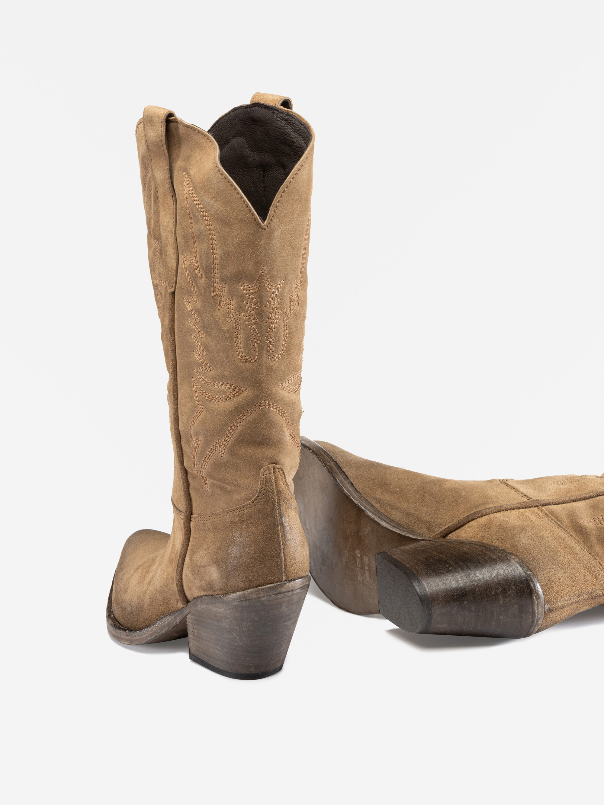 Ein Paar hellbraune Cowboystiefel aus Wildleder mit Ziernähten, einer aufrecht stehend, der andere auf der Seite liegend, beide mit gestapelten Holzabsätzen, vor weißem Hintergrund.