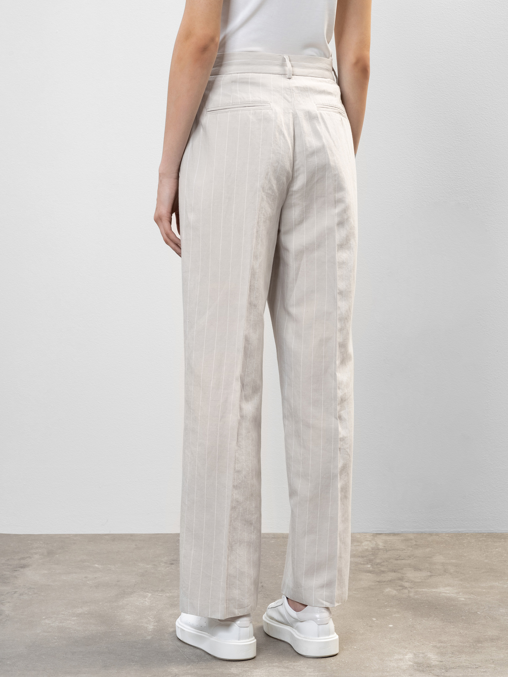 Eine Person steht mit dem Gesicht zur Wand, bekleidet mit einer hellbeigen, gestreiften Hose mit weitem Bein, einem weißen Oberteil und weißen Plateau-Turnschuhen, auf einem grauen Betonboden vor einer schlichten, hellen Wand.