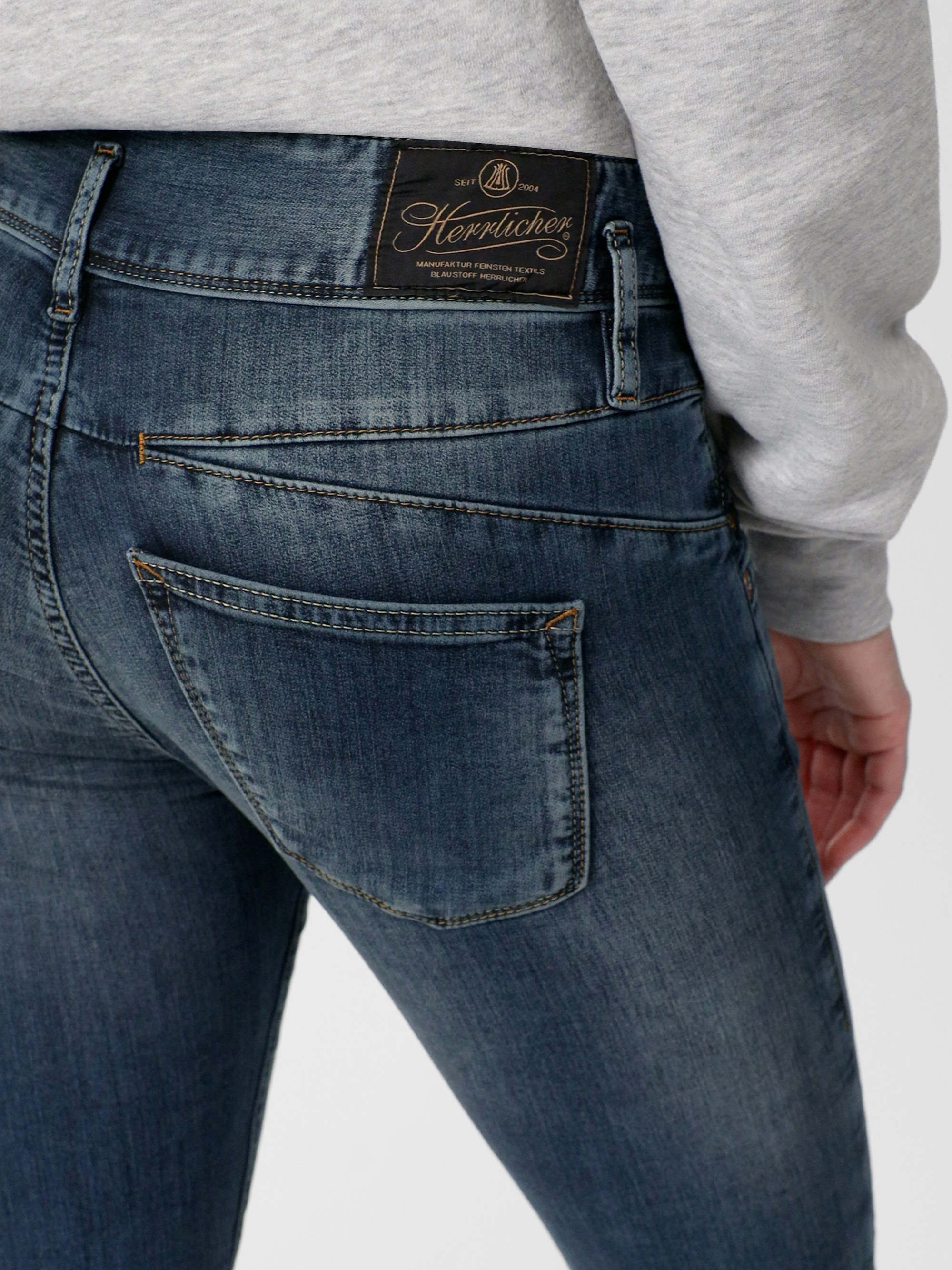 Nahaufnahme einer Person, die eine blaue Denim-Jeans und ein hellgraues Sweatshirt trägt, wobei die Gesäßtasche und ein schwarzer Markenaufnäher aus Leder am Bund zu sehen sind.