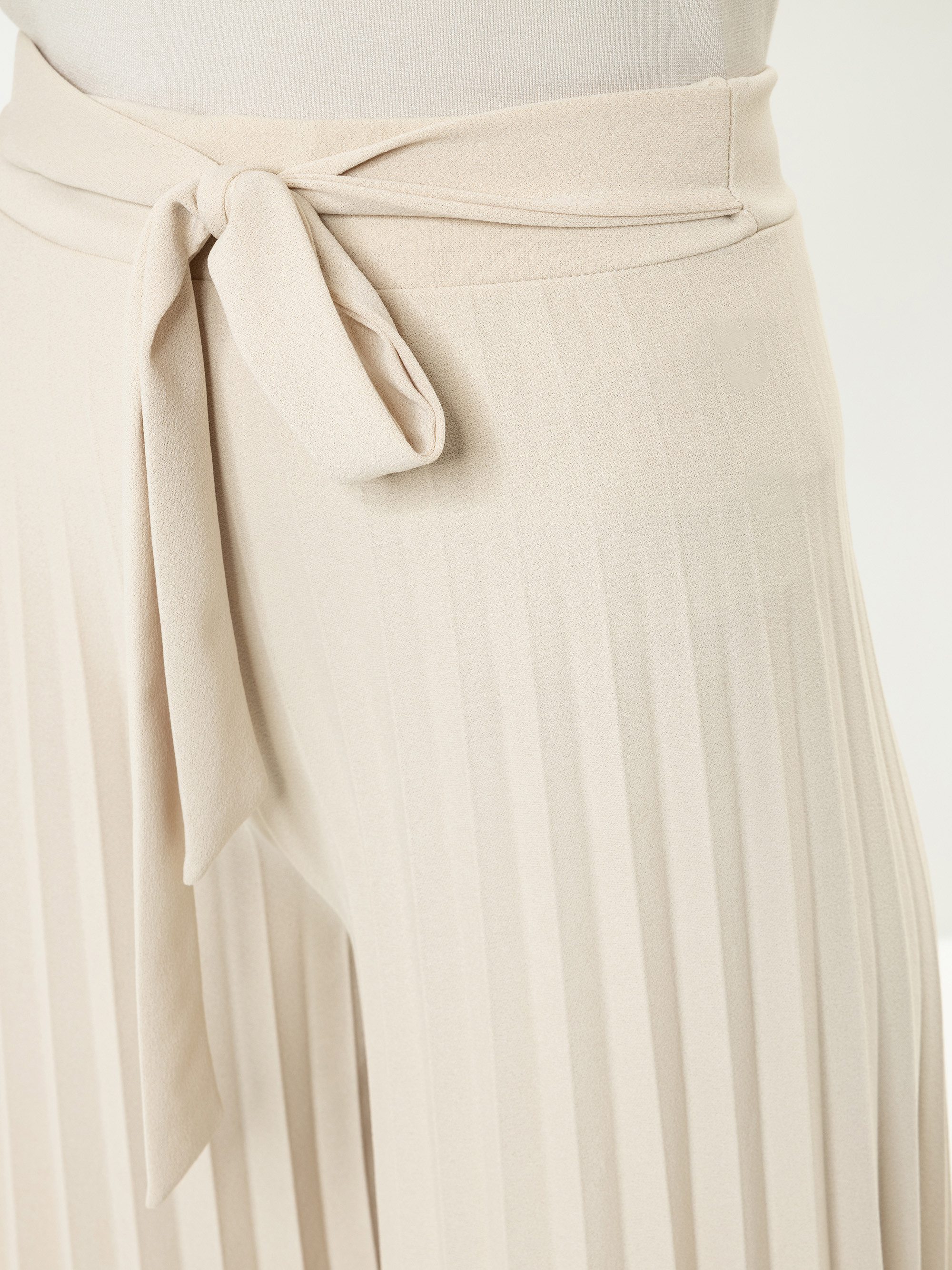 Nahaufnahme eines cremefarbenen Faltenrocks oder einer Hose mit einem Stoffgürtel, der in der Taille verknotet ist. Die Textur und die vertikalen Falten sind deutlich sichtbar.