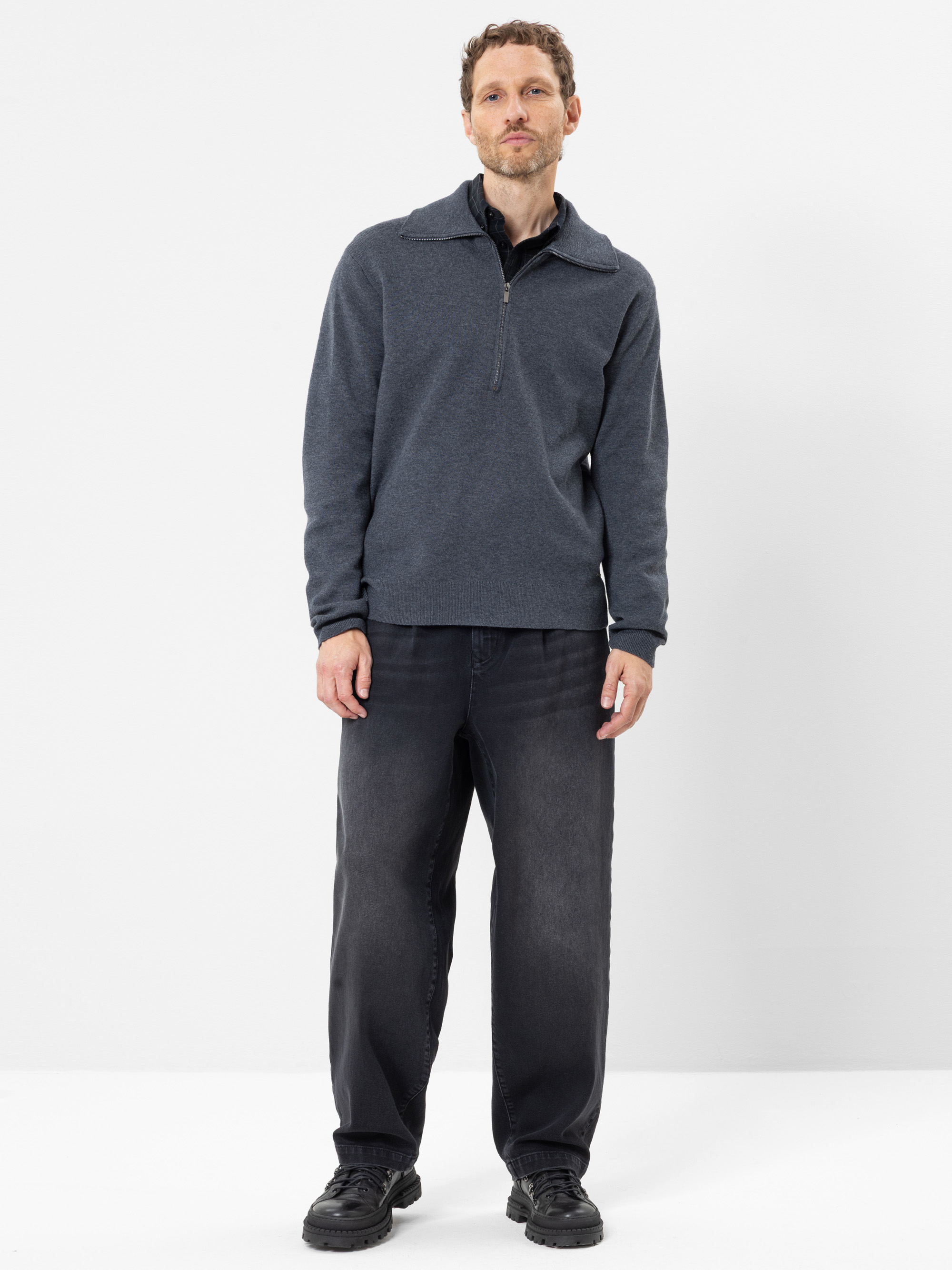 Vor einem schlichten weißen Hintergrund steht ein Mann, der einen dunkelgrauen Pullover mit Viertelreißverschluss, schwarze Jeans in lockerer Passform und schwarze Turnschuhe trägt. Er hat kurzes, lockiges Haar und einen gepflegten Bart, sein Gesichtsa