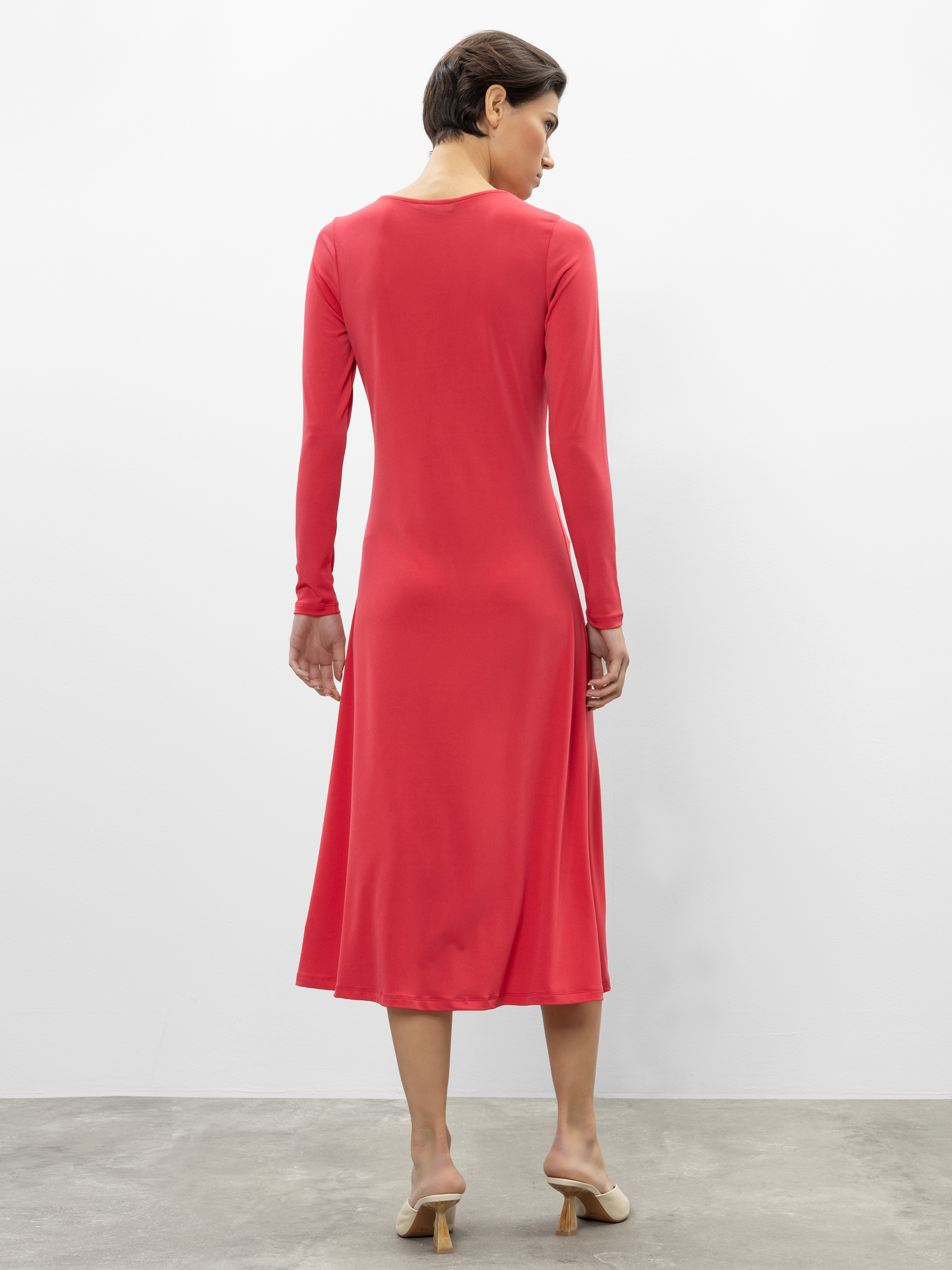 Eine Frau in einem langärmeligen, korallenroten Midikleid steht von der Kamera abgewandt. Sie hat kurzes braunes Haar und trägt beigefarbene Pantoletten mit Absatz. Der Hintergrund ist eine schlichte weiße Wand mit einem grauen Boden.