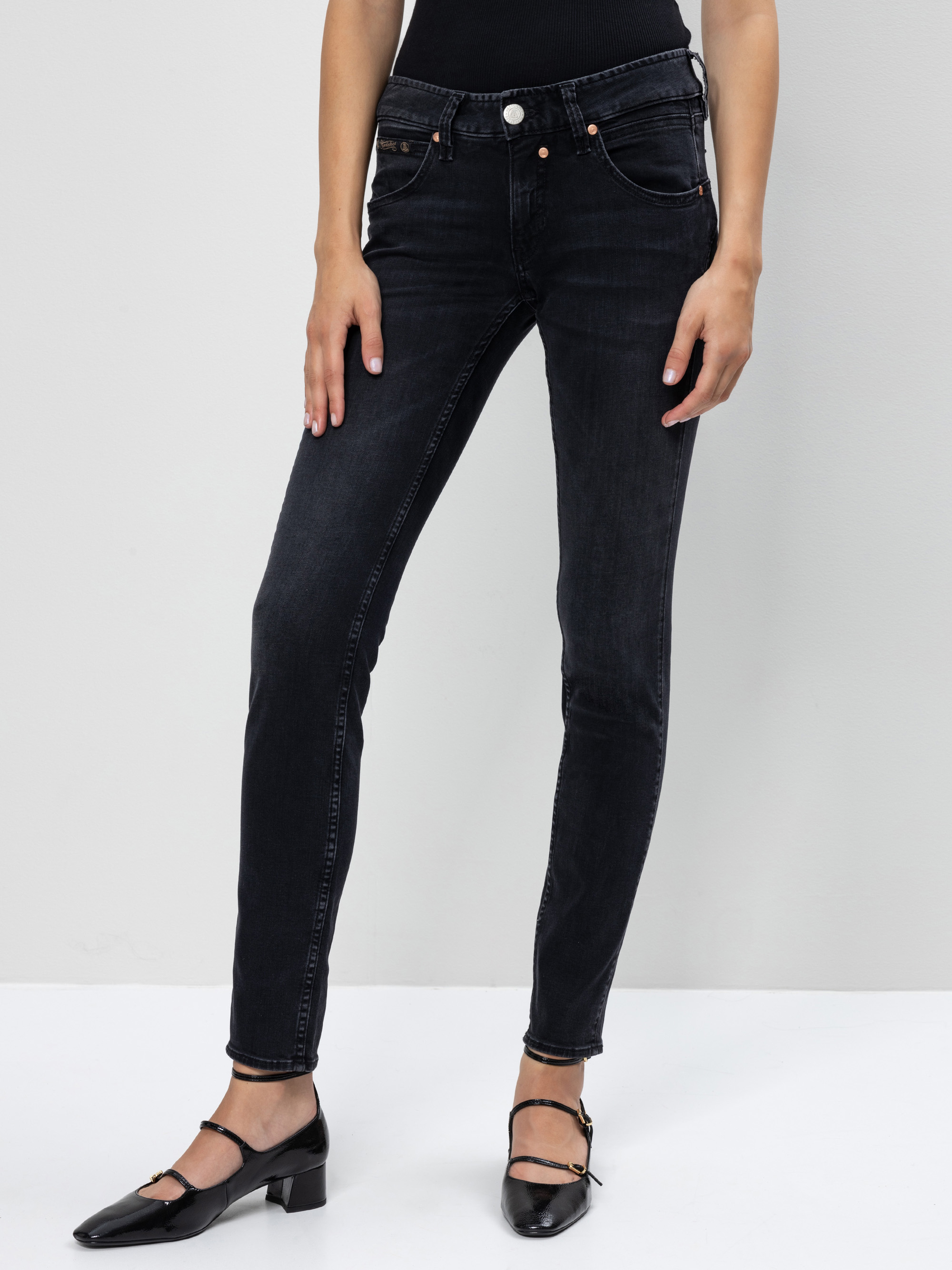 Person in schwarzer Jeans und schwarzen Mary-Jane-Schuhen mit Absatz, stehend vor einem schlichten hellgrauen Hintergrund. Die obere Hälfte des Körpers der Person ist nicht sichtbar.