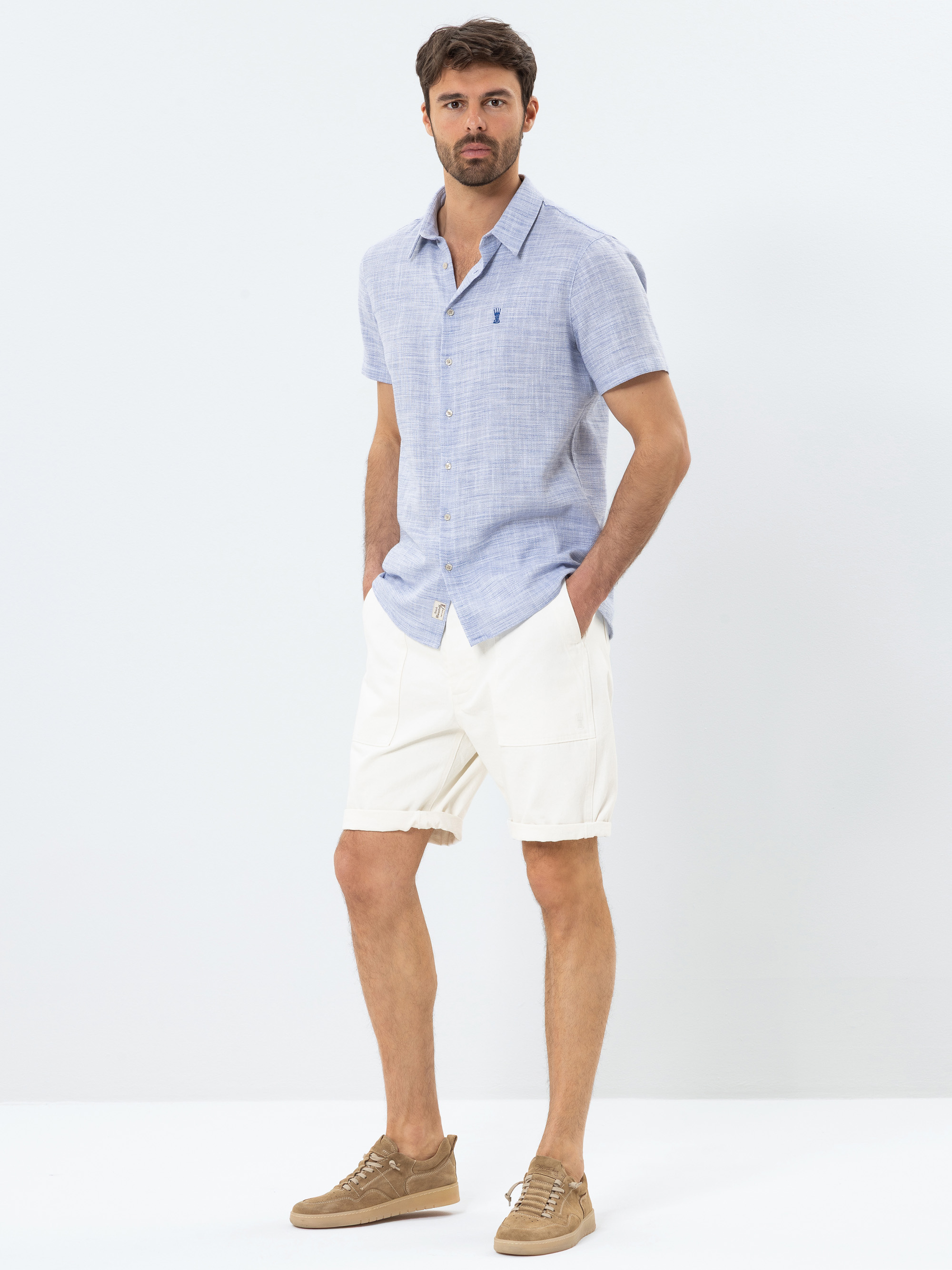 Ein Mann steht mit den Händen in den Taschen, trägt ein hellblaues, kurzärmeliges Button-up-Hemd, weiße Shorts und braune Turnschuhe vor einem schlichten, hellen Hintergrund.