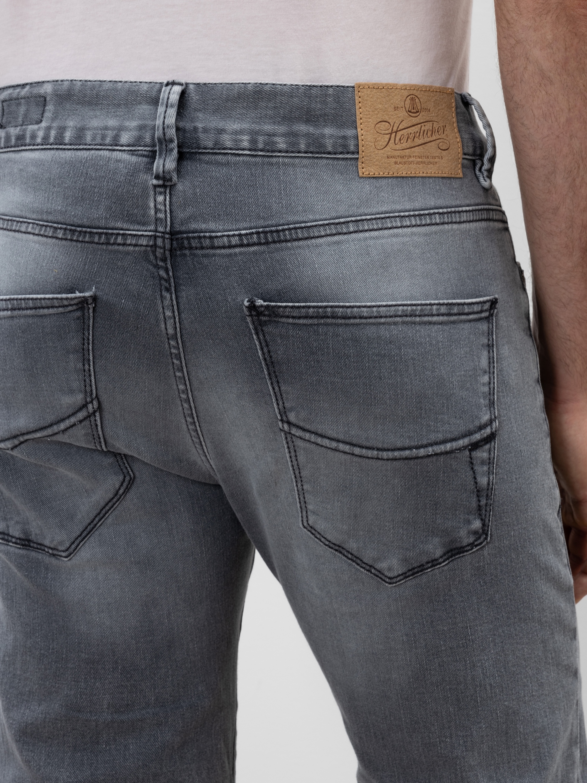 Nahaufnahme des Rückens einer Person, die eine graue Jeans mit zwei Gesäßtaschen und einem braunen Markenaufnäher aus Leder am Bund trägt. Die Person trägt außerdem ein helles Hemd, das in die Hose gesteckt ist.