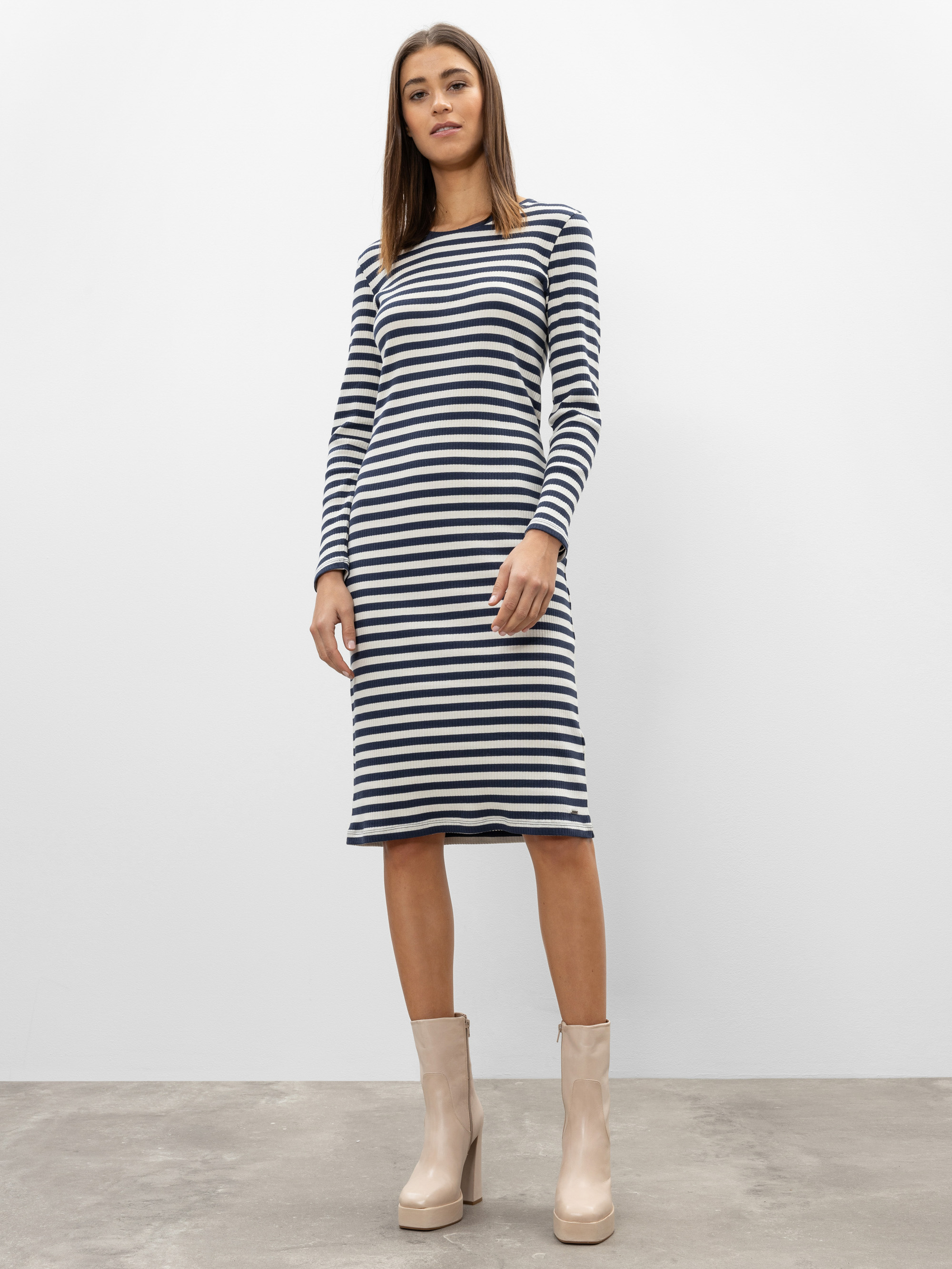 Eine Frau steht vor einem schlichten Hintergrund und trägt ein schwarz-weiß gestreiftes, knielanges Kleid und beigefarbene Plateaustiefel.