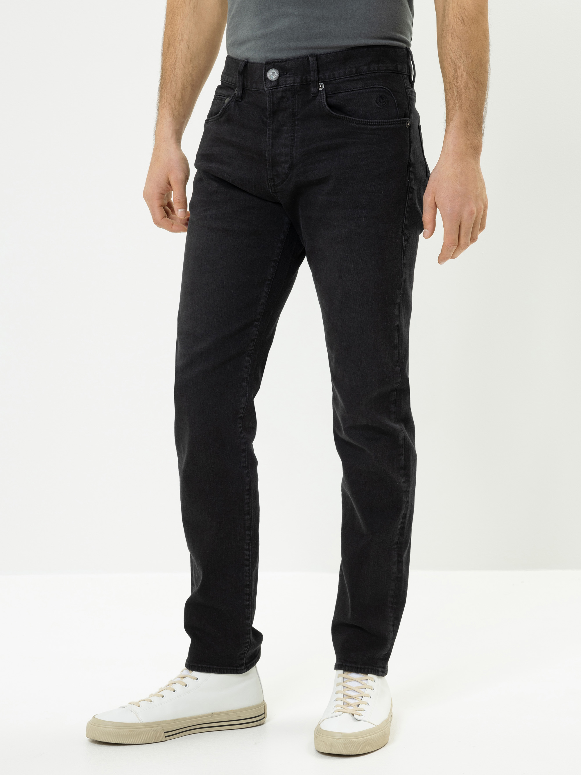 Ein Mann, der schwarze Jeans, ein dunkelgraues T-Shirt und weiße Turnschuhe trägt, steht vor einem schlichten weißen Hintergrund. Sein Kopf ist auf dem Bild nicht zu sehen.