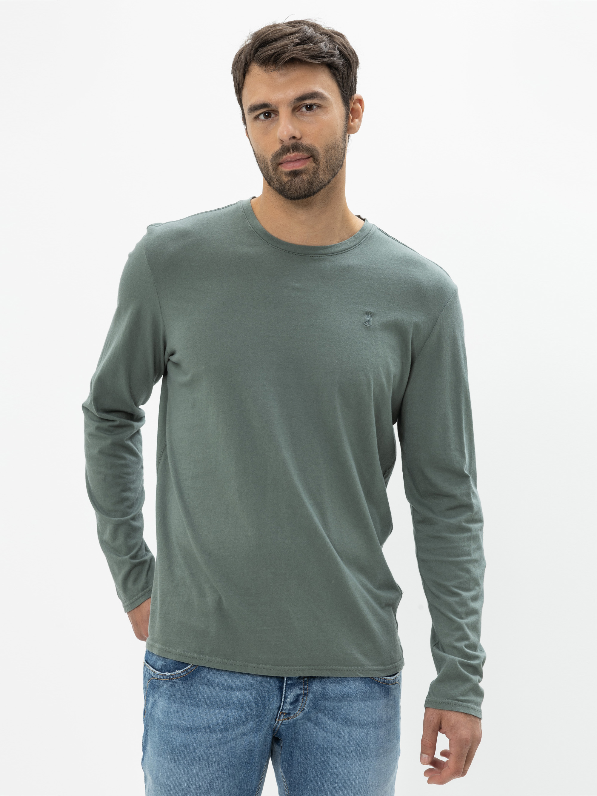 Ein Mann mit kurzen dunklen Haaren und einem Bart trägt ein salbeigrünes, langärmeliges Hemd und hellblaue Jeans und steht mit einer Hand in der Tasche vor einem schlichten weißen Hintergrund.