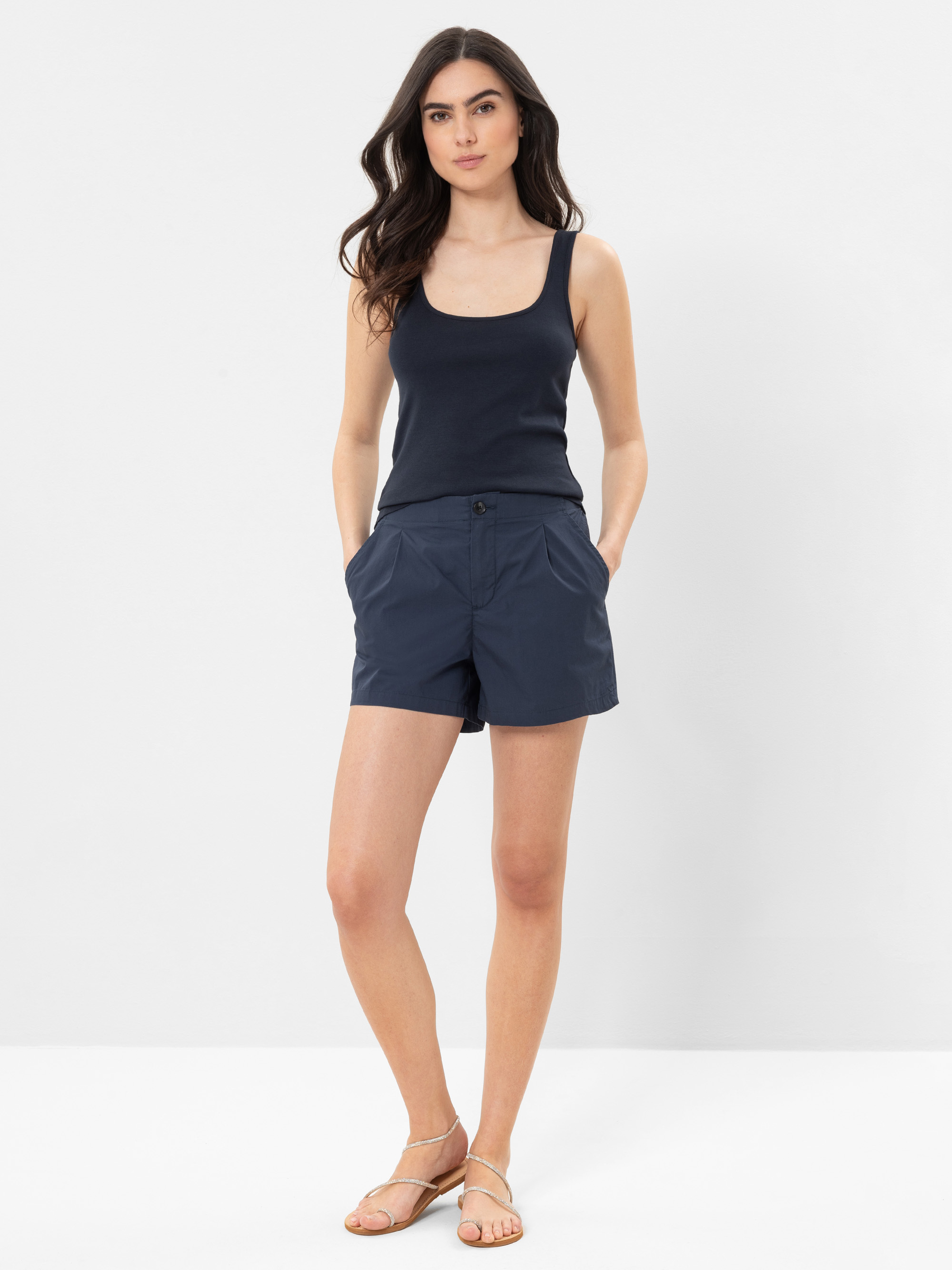Eine Frau steht vor einem schlichten weißen Hintergrund und trägt ein marineblaues Tanktop, marineblaue Shorts und beigefarbene Sandalen. Sie hat langes dunkles Haar und ihre Hände stecken in den Taschen.
