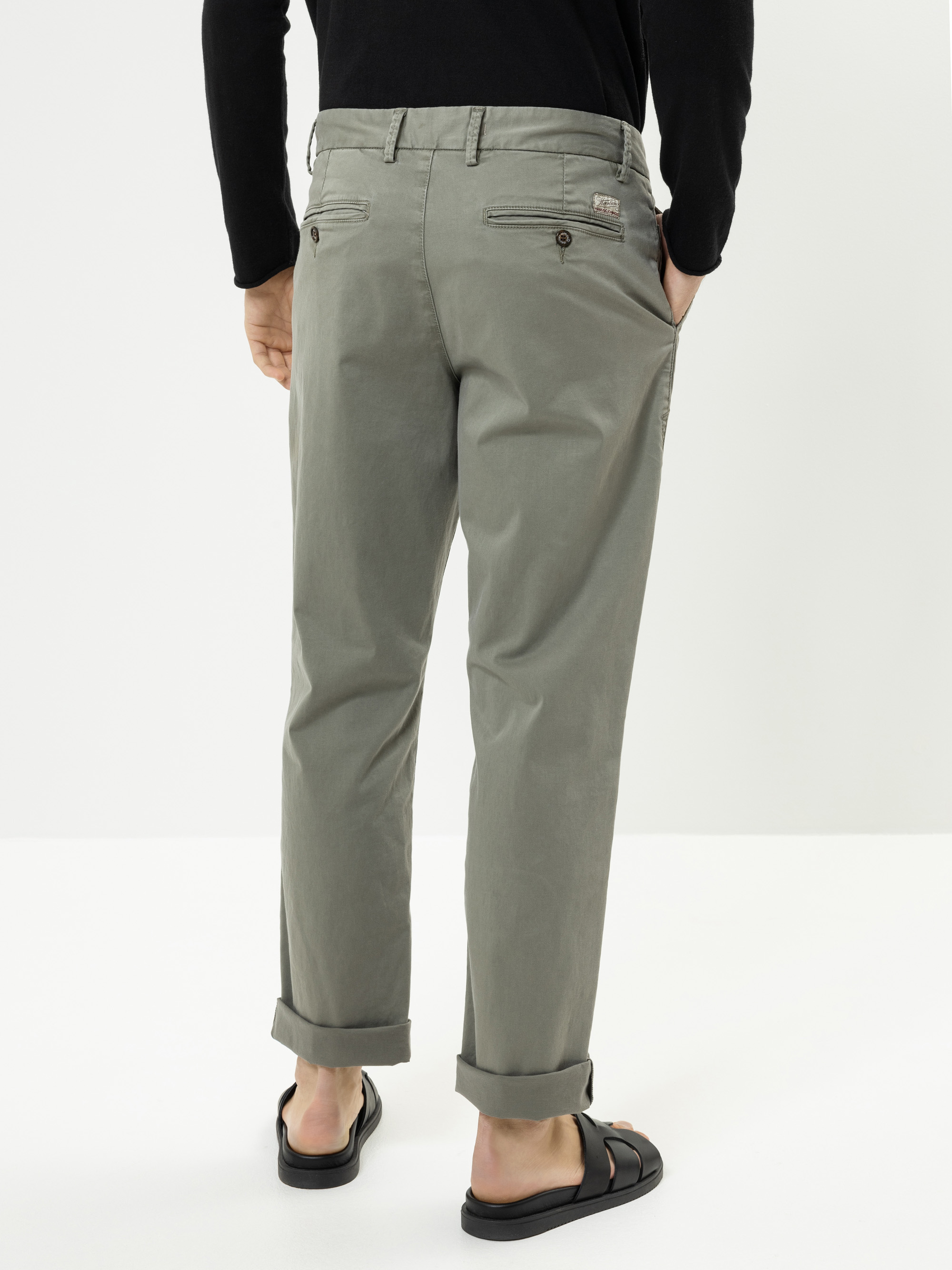 Eine Person, die eine salbeigrüne Hose mit Bündchen, ein schwarzes langärmeliges Oberteil und schwarze Sandalen trägt, steht vor einem schlichten weißen Hintergrund, von hinten gesehen.