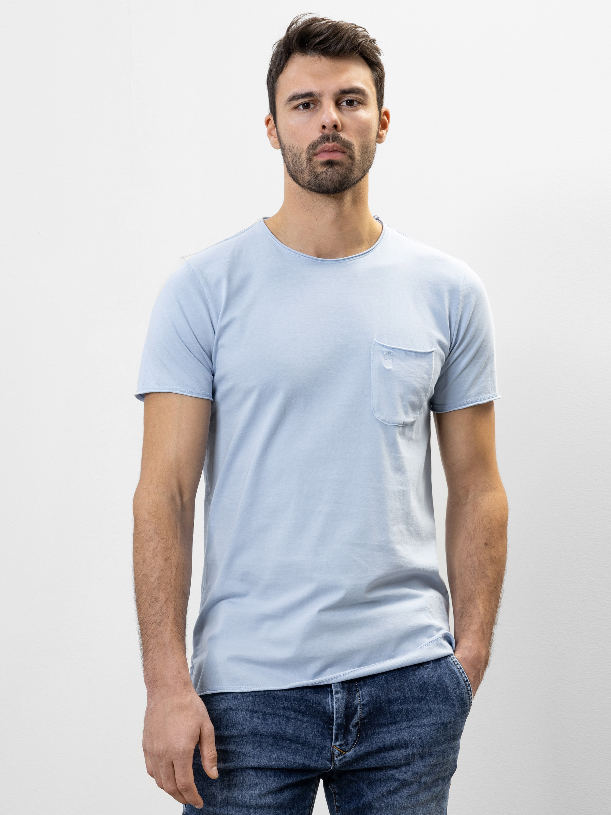 Ein Mann mit kurzen dunklen Haaren und einem Bart steht vor einem schlichten weißen Hintergrund. Er trägt ein hellblaues T-Shirt mit einer Brusttasche und eine blaue Jeans. Seine Hände liegen entspannt an den Seiten, und er hat einen neutralen Gesichts