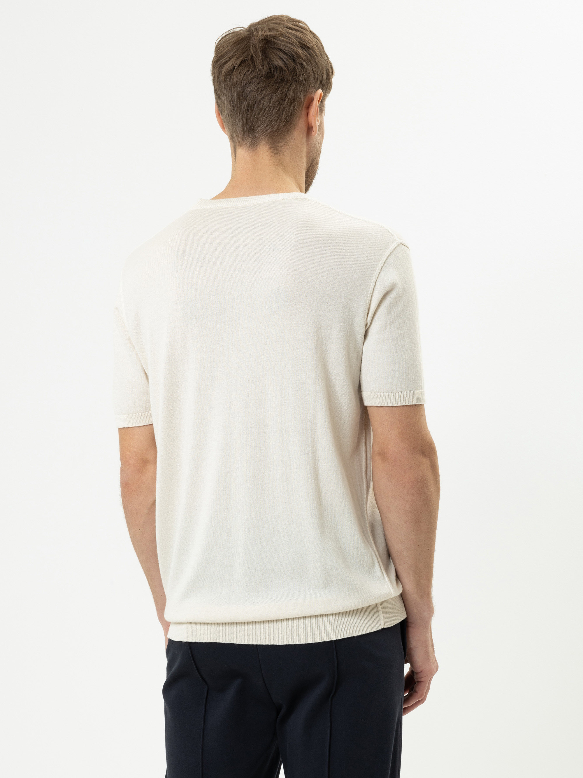 Ein Mann mit kurzen braunen Haaren blickt von der Kamera weg und trägt ein kurzärmeliges, hellbeiges Strickhemd und eine dunkle Hose vor einem schlichten weißen Hintergrund.