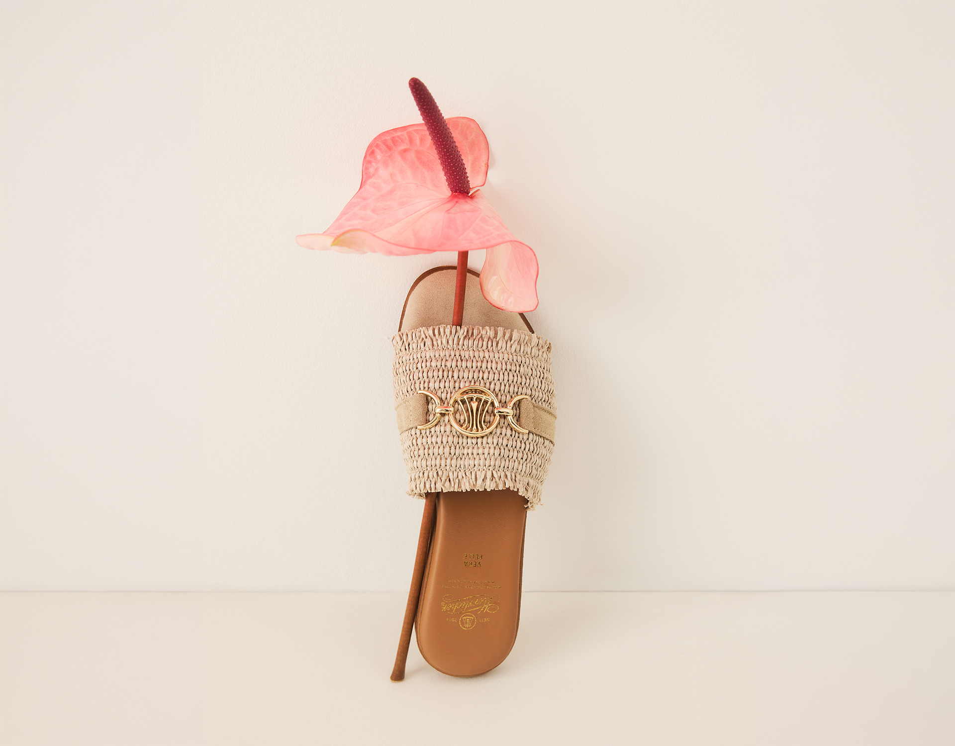 Eine beigefarbene gewebte Sandale mit goldenen Akzenten steht aufrecht, mit einer rosafarbenen Anthurienblüte und ihrer roten Spadix, die durch die Öffnung der Sandale gesteckt wird, vor einem schlichten, cremefarbenen Hintergrund.