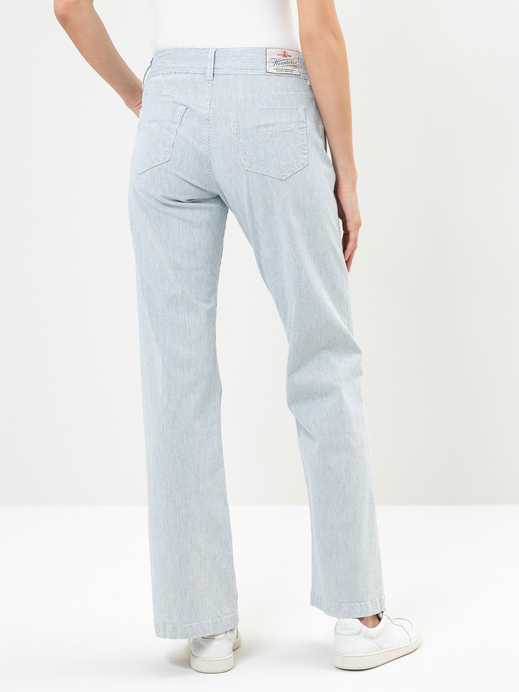 Eine Person steht mit dem Gesicht zur Wand, trägt hellblaue Jeans mit hoher Taille, ein weißes ärmelloses Oberteil und weiße Turnschuhe vor einem schlichten weißen Hintergrund.