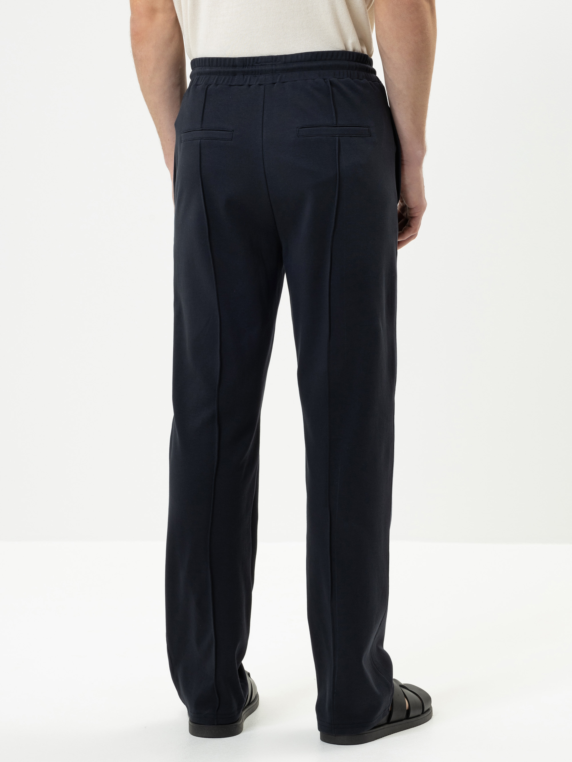 Eine Person steht mit dem Gesicht zur Wand, trägt ein beiges Hemd, eine dunkle marineblaue Hose mit Leistentaschen und schwarze Sandalen vor einem schlichten weißen Hintergrund.