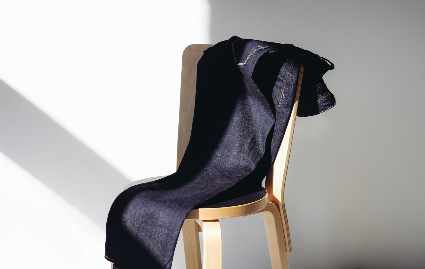 Eine dunkelblaue Jeans ist über einen hellen Holzstuhl drapiert, während das Sonnenlicht Schatten auf eine weiße Wand im Hintergrund wirft.