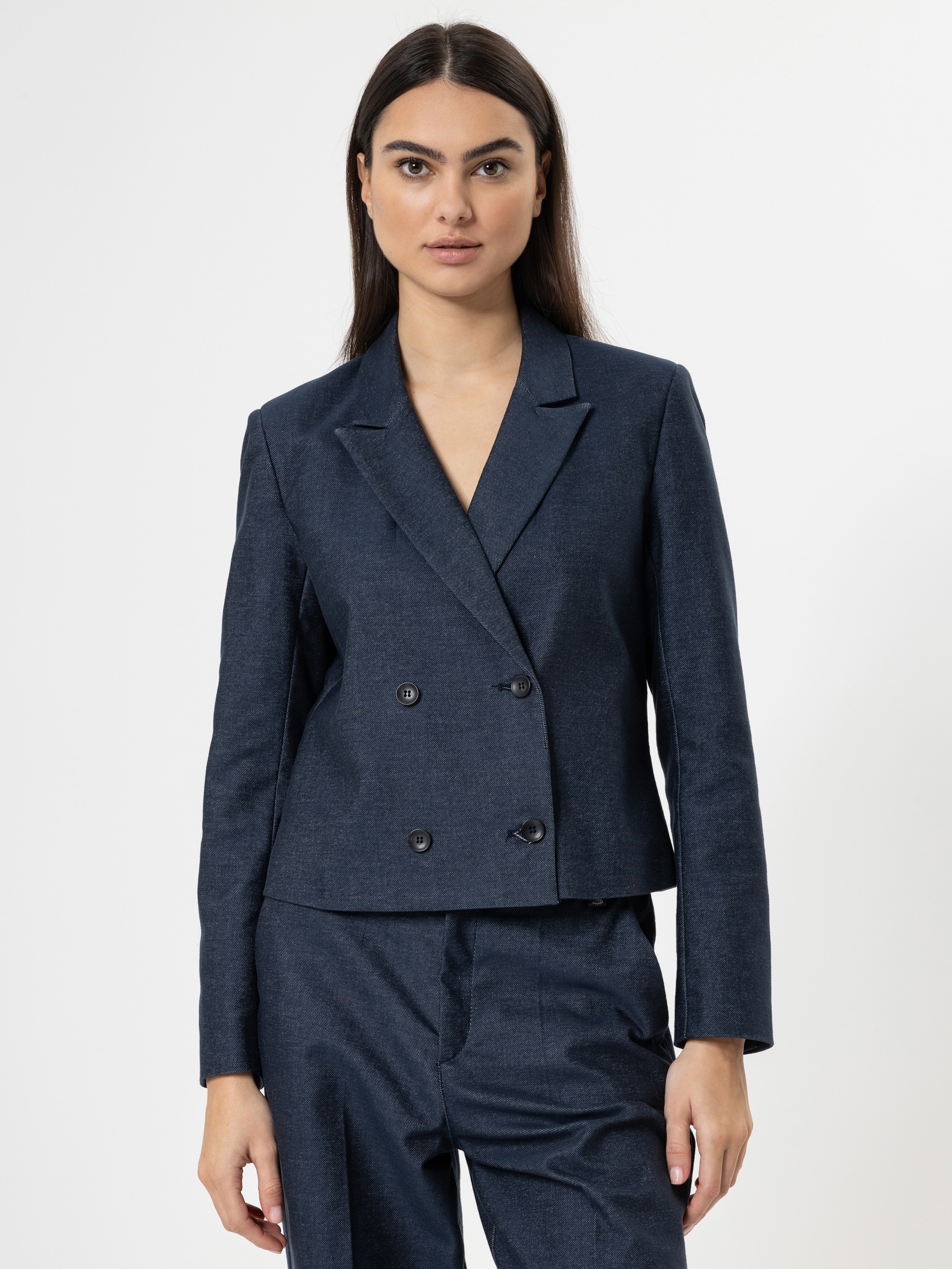 Eine Frau mit glattem dunklem Haar trägt einen marineblauen Zweireiher mit langen Ärmeln und einer passenden Hose vor einem schlichten weißen Hintergrund.