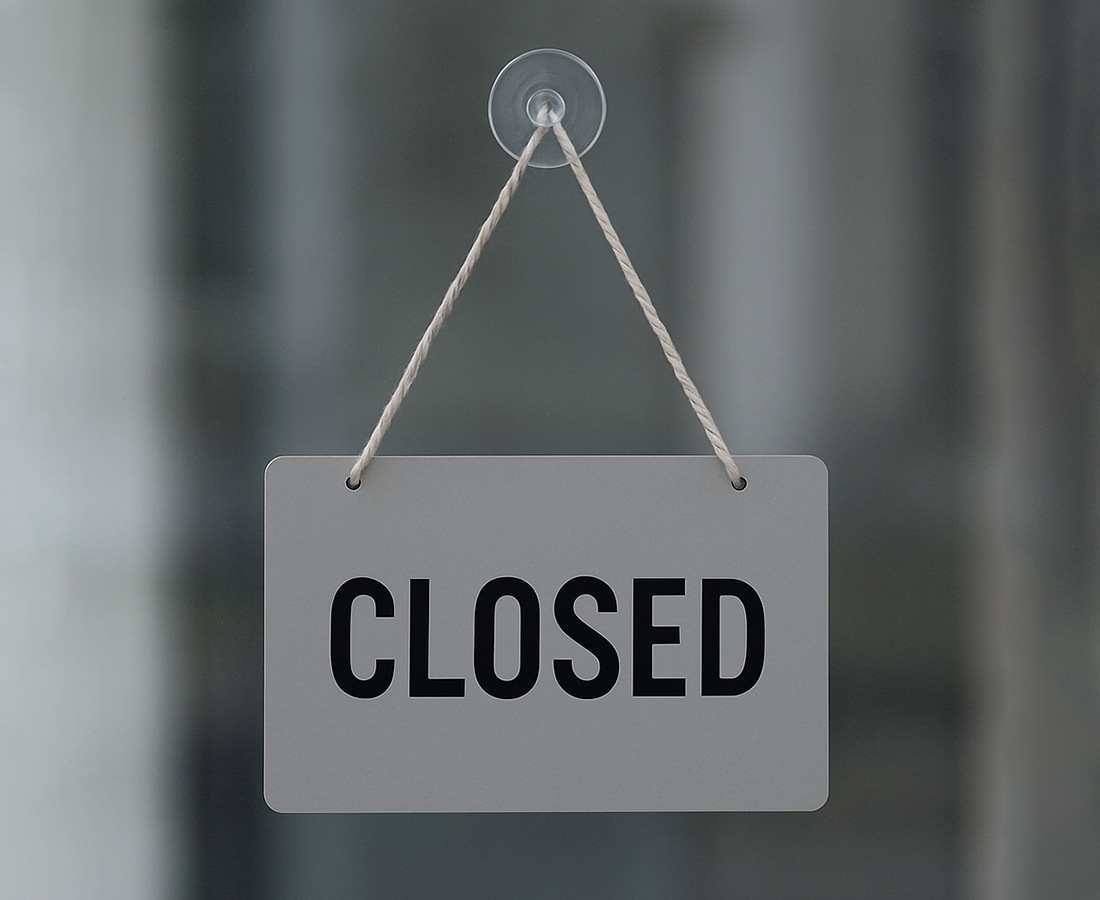Ein graues Schild mit dem Wort GESCHLOSSEN in fetten schwarzen Buchstaben hängt an einer weißen Schnur, die an einem Saugnapf an einer Glastür oder einem Fenster befestigt ist. Der Hintergrund erscheint unscharf.