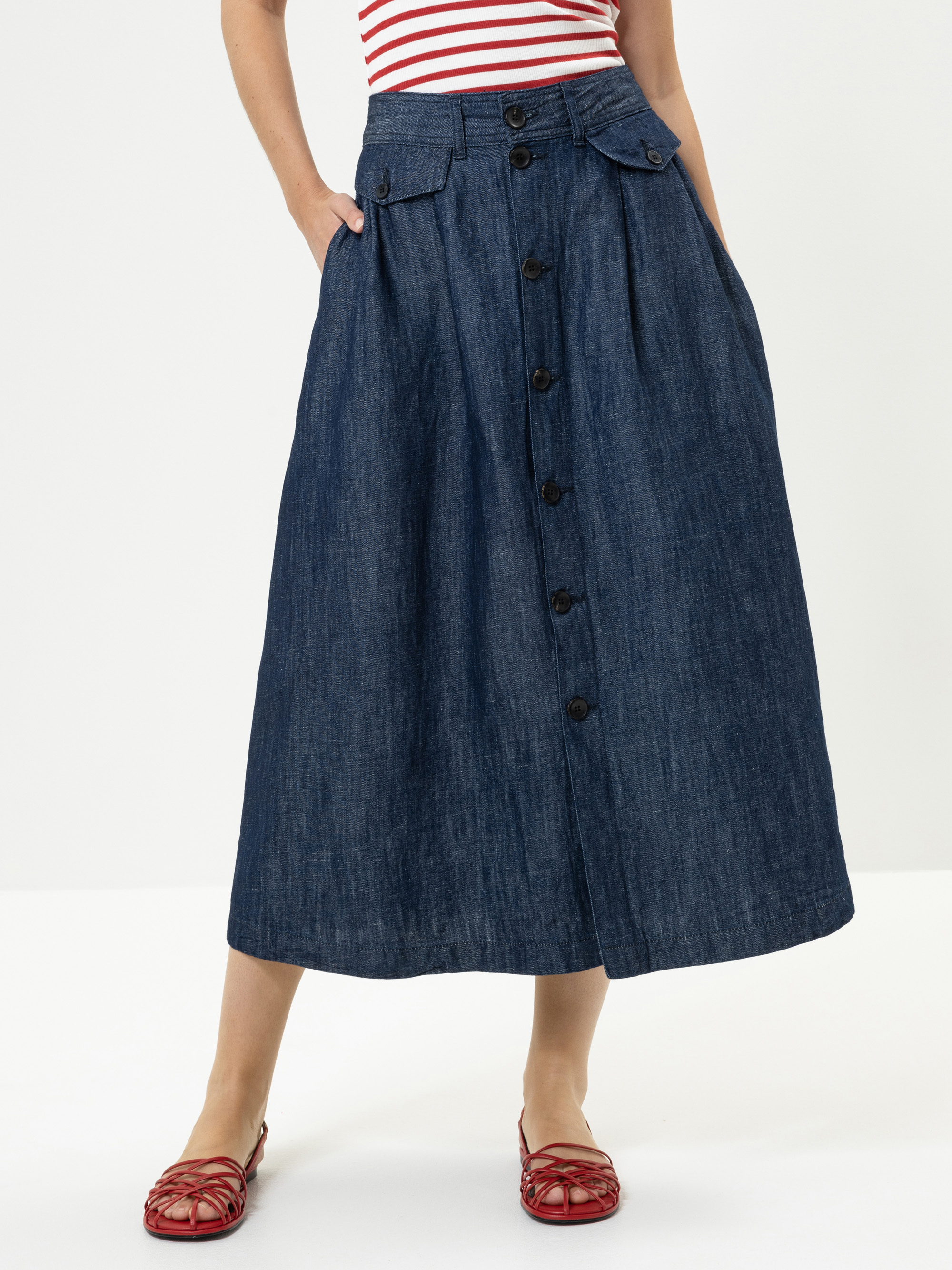 Eine Person, die einen hochtaillierten Jeans-Midirock mit Knöpfen und Taschen trägt, dazu ein rot-weiß gestreiftes Oberteil und rote Webschuhe, steht vor einem schlichten weißen Hintergrund.