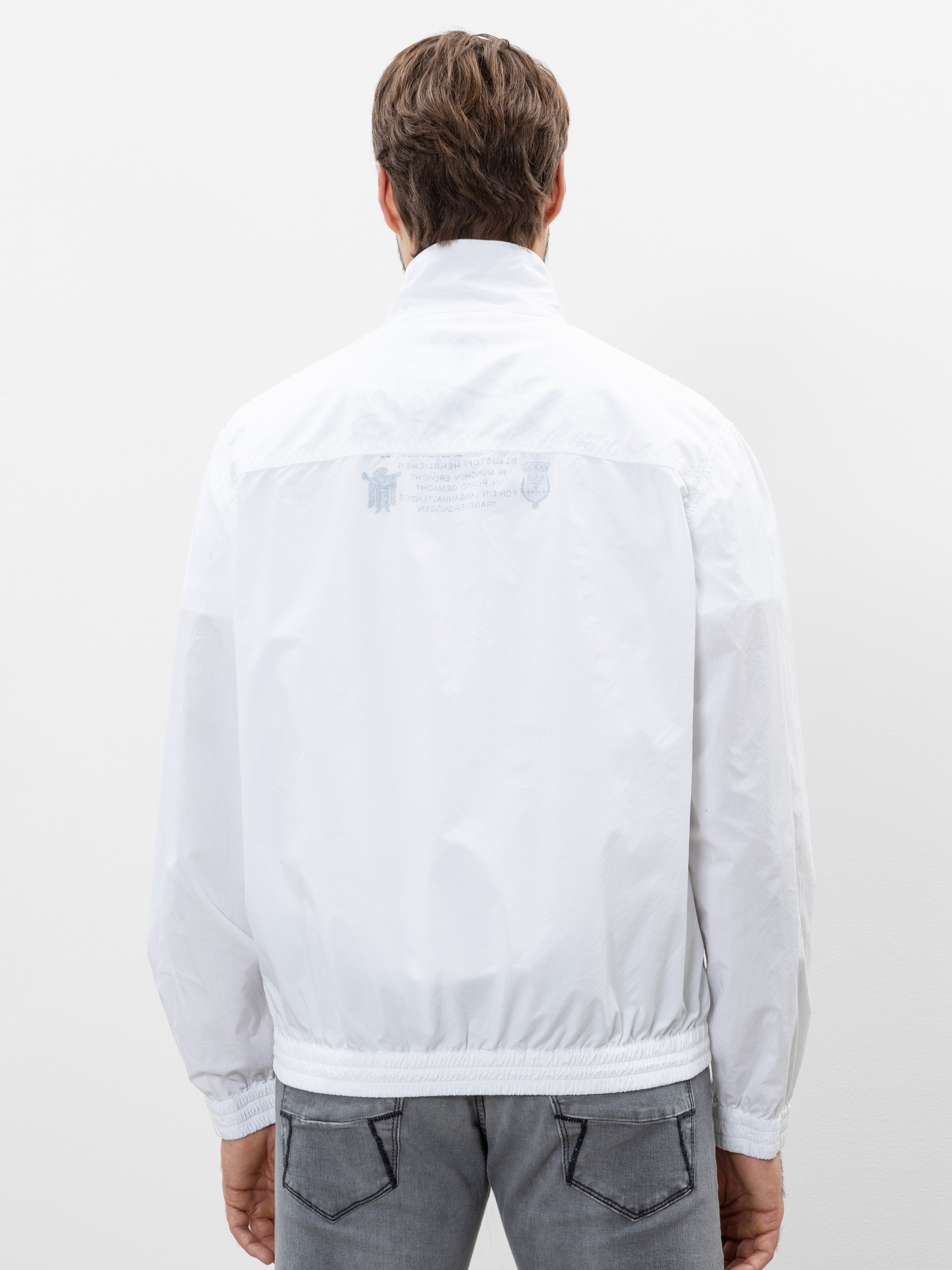 Eine Person mit kurzen braunen Haaren trägt eine weiße Jacke und graue Jeans und blickt von der Kamera weg auf einen schlichten weißen Hintergrund.