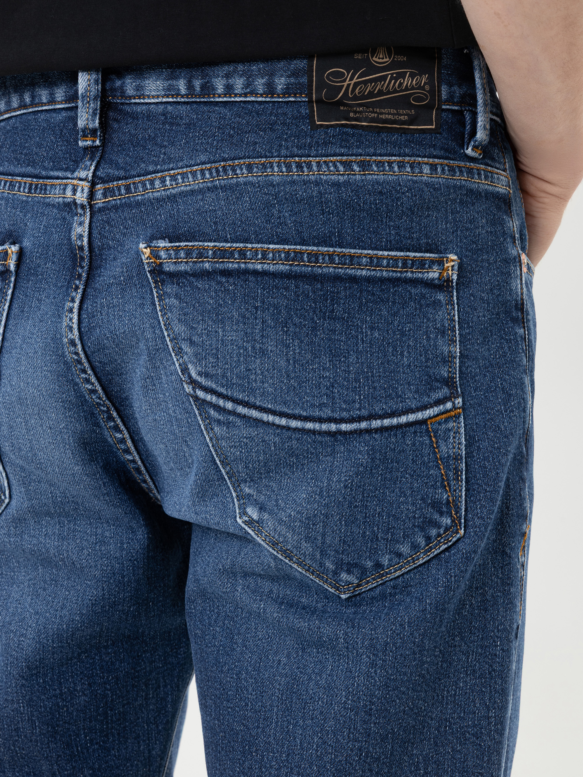 Nahaufnahme des Rückens einer Person, die blaue Jeans trägt, mit sichtbaren Nähten und einem schwarzen Leder-Aufnäher der Marke „Herrlicher“ über der rechten Tasche.