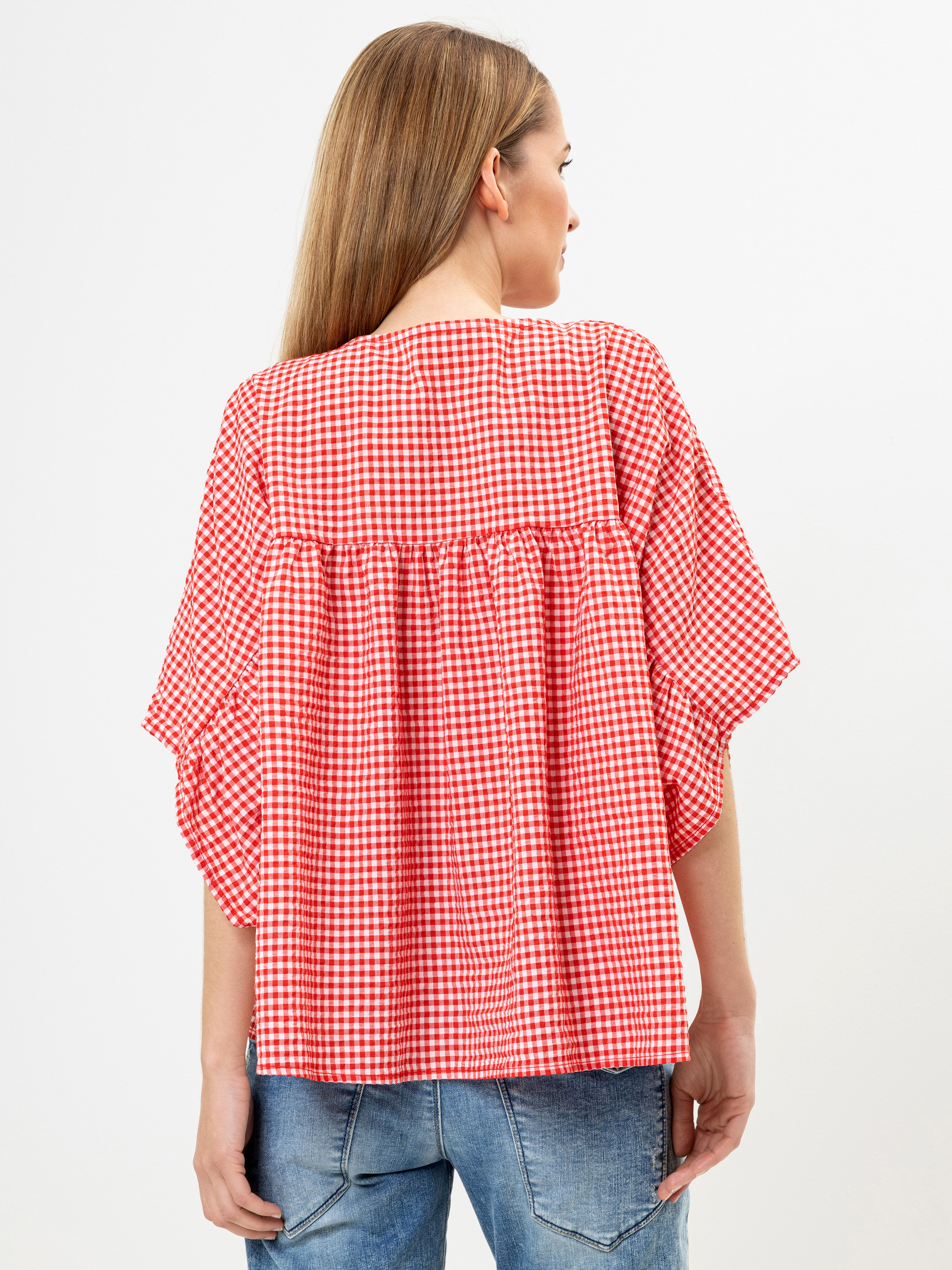 Eine Frau mit langem, glattem Haar wird von hinten gezeigt. Sie trägt eine locker sitzende, rot-weiß karierte Bluse mit kurzen Ärmeln und blaue Jeans. Der Hintergrund ist einfarbig weiß.